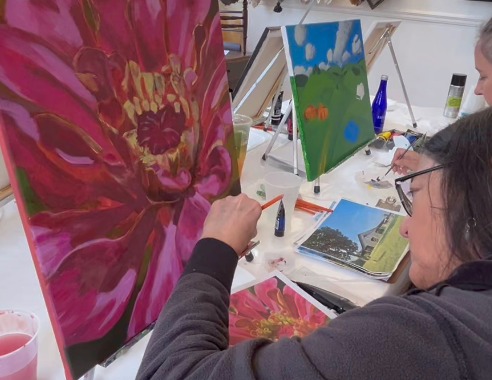 People painting on canvases at an indoor art class, with paintings of flowers and landscapes on easels and tables around them.