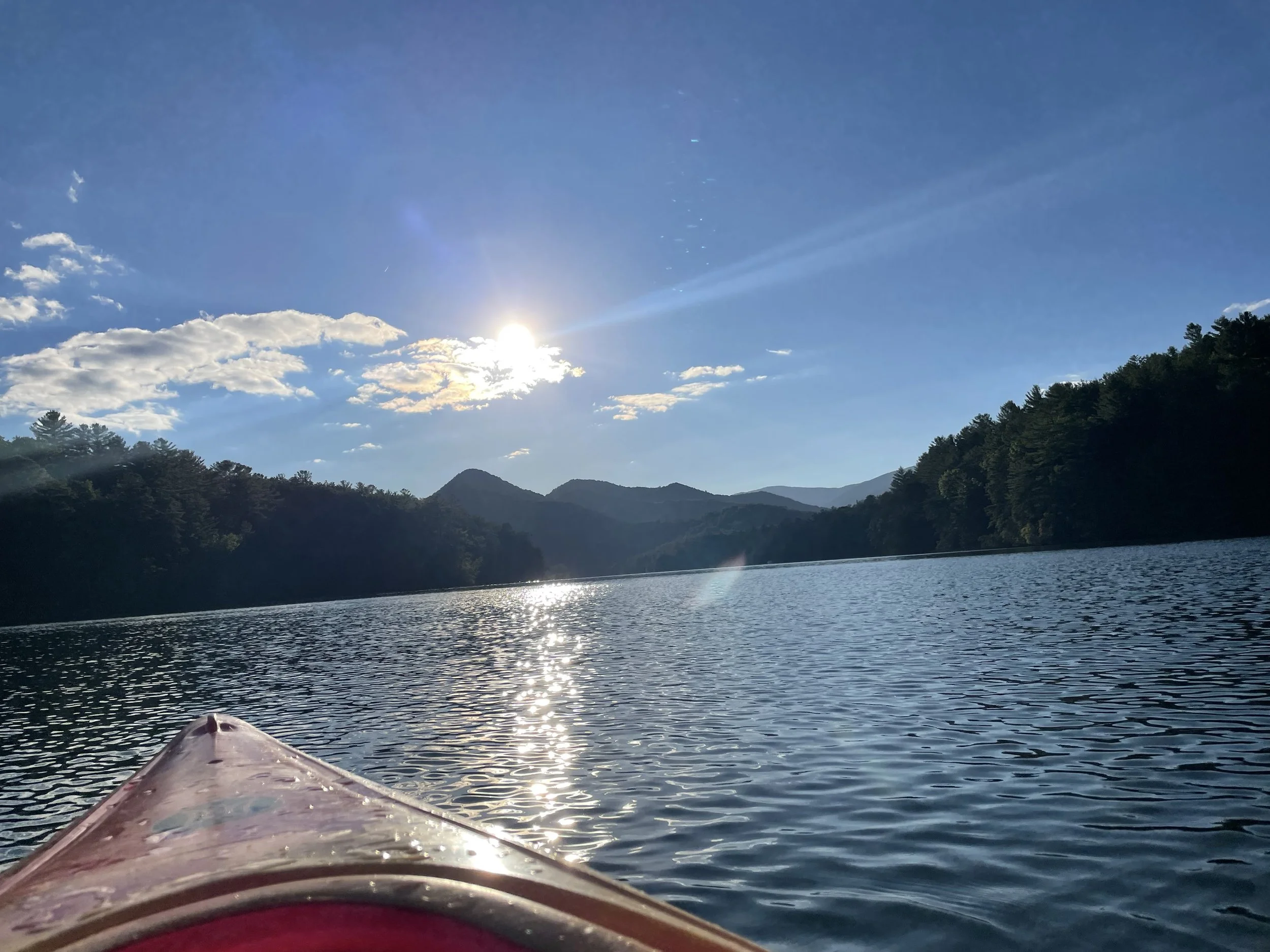 A kayak on a calm lake surrounded by forested hills and mountains with a bright sun and blue sky with some clouds.