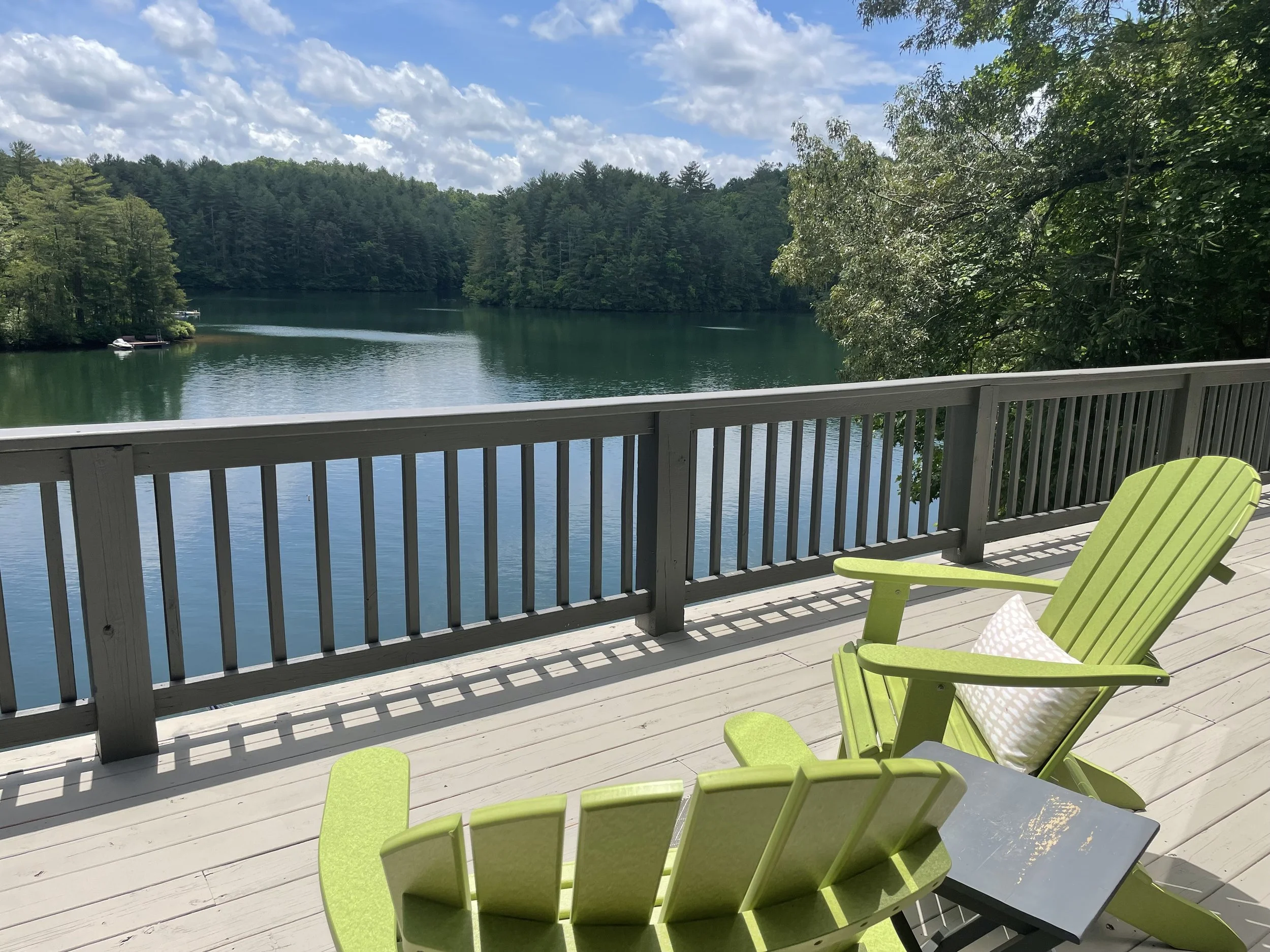View of a lake with a forested shoreline, on a wooden deck with two green Adirondack chairs and a small side table.