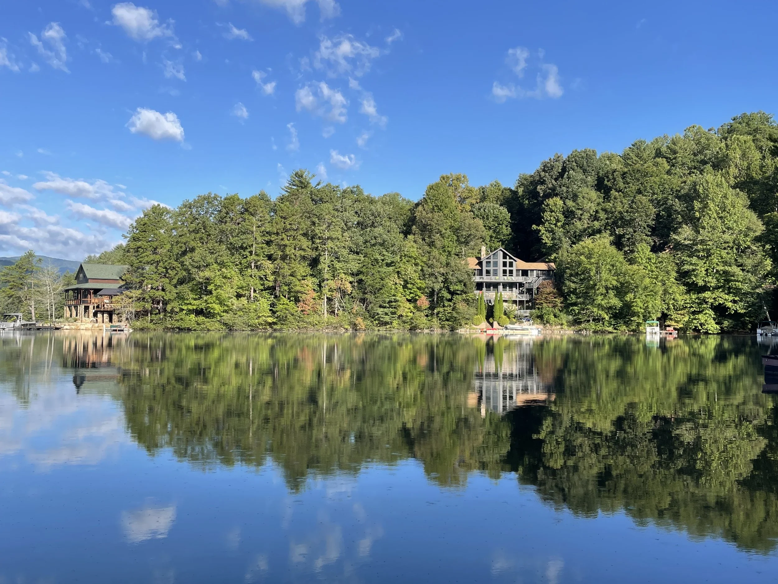A peaceful lakeside scene with calm water reflecting green trees, houses, and boats, under a bright blue sky with scattered clouds.