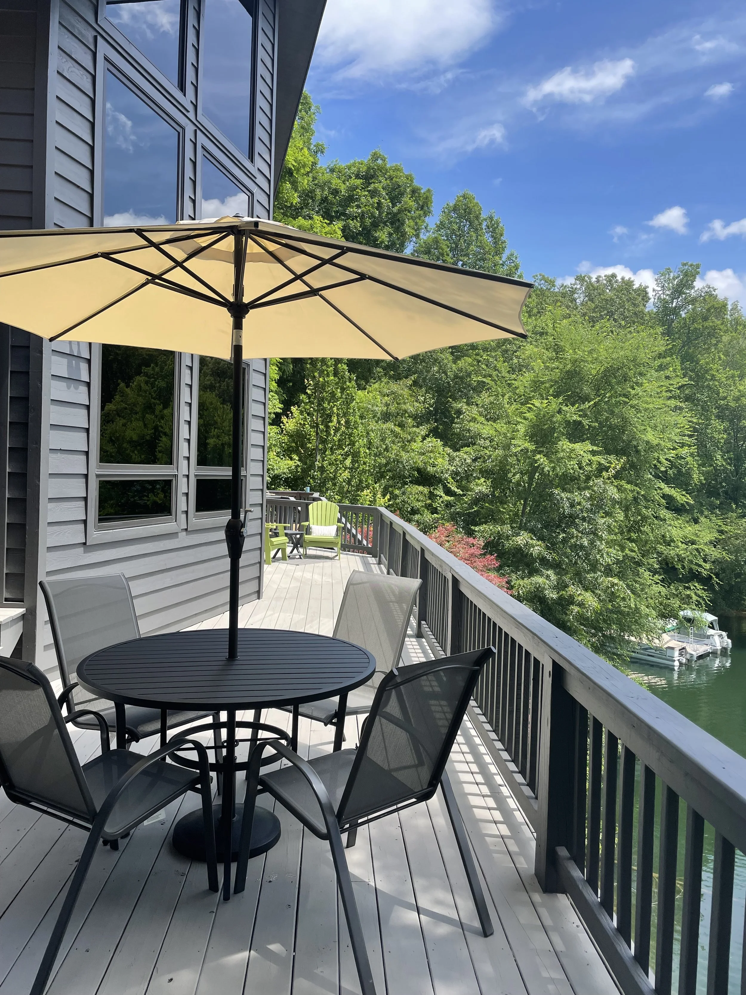 Outdoor balcony with a black round table, four chairs, a beige umbrella, and green chairs at the end. Trees and a boat on the water are visible in the background.