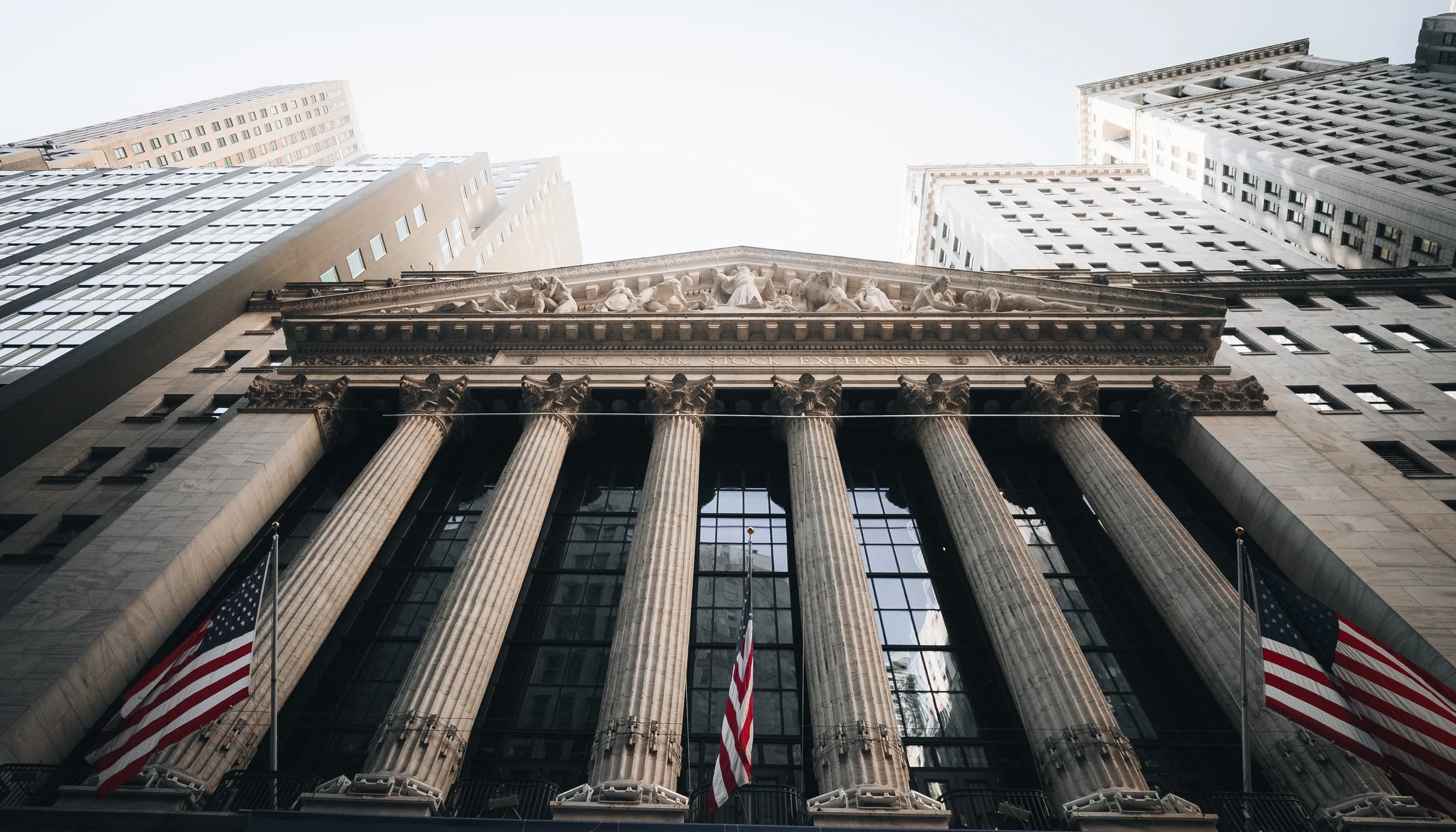 Low-angle view of the New York Stock Exchange building with tall skyscrapers in the background and American flags in the foreground.