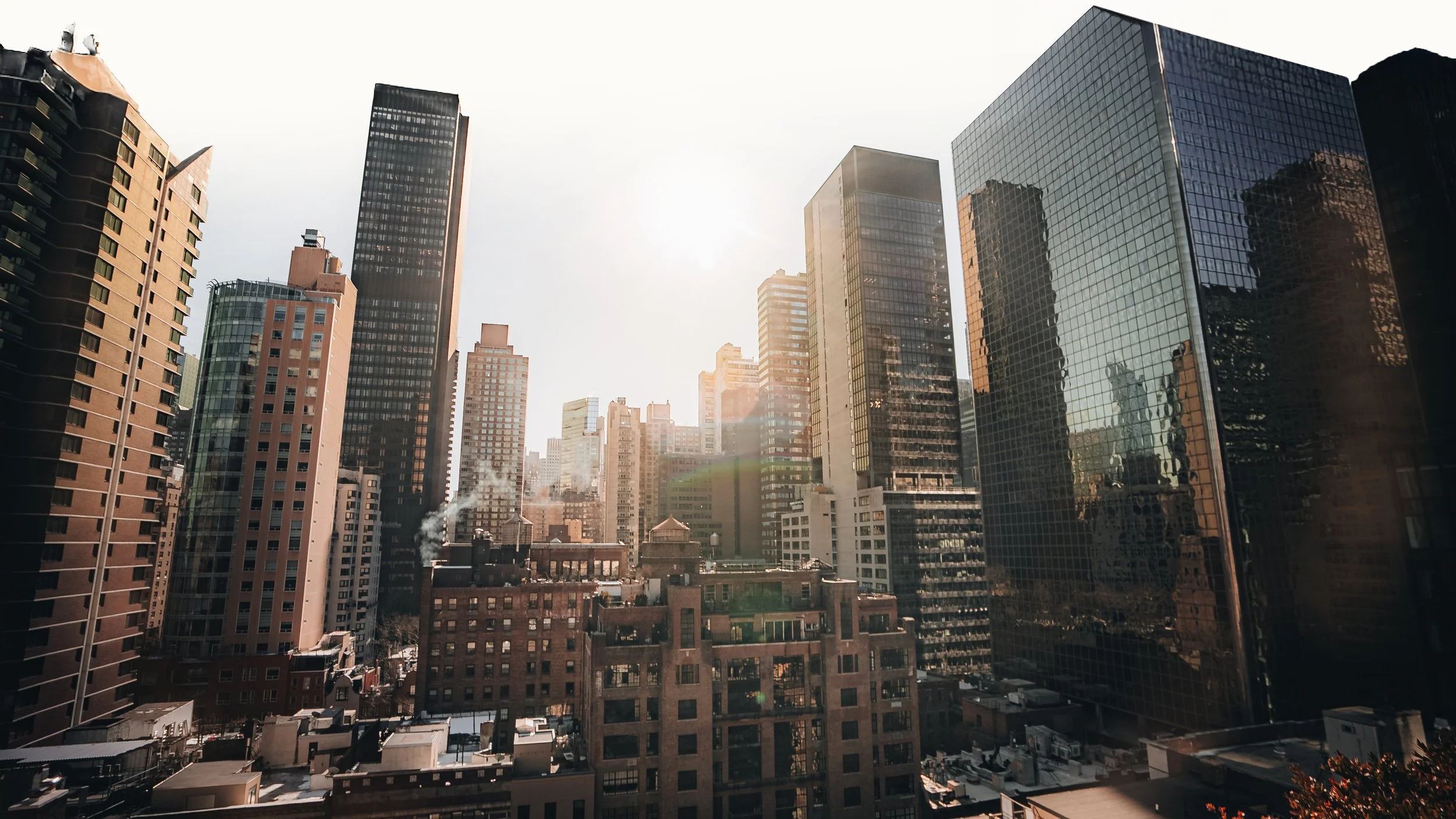 Cityscape of tall buildings during sunset with glass and brick skyscrapers and a bright sky.