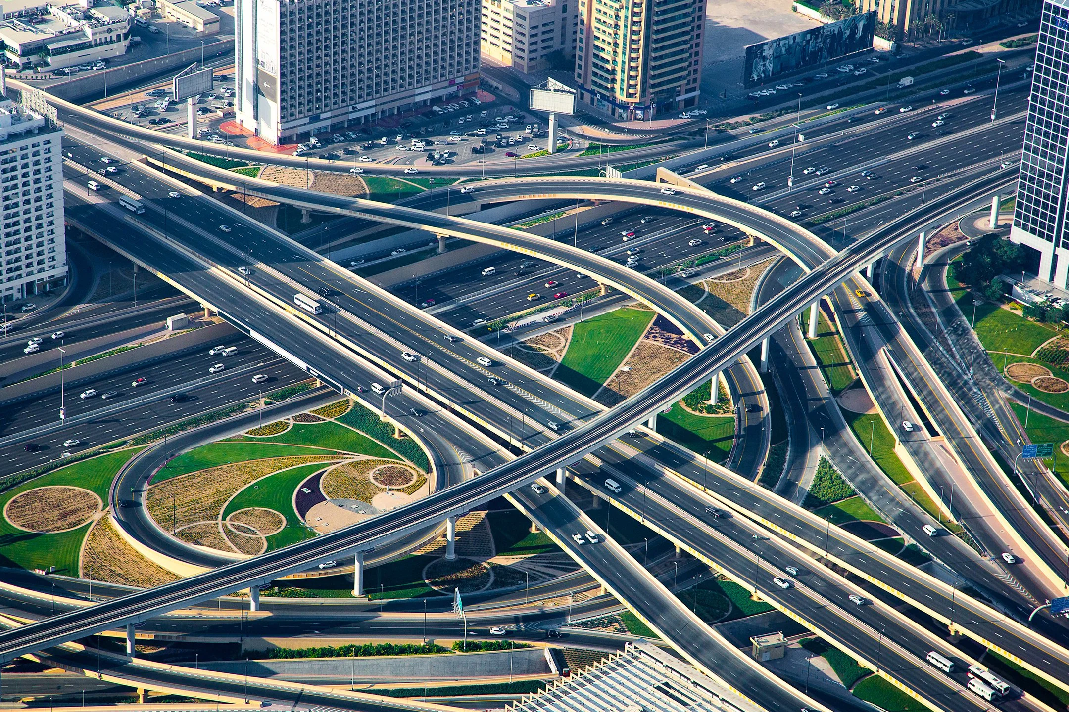An aerial view of a multi-level highway interchange with multiple overpasses and ramps, surrounded by tall commercial buildings and some green landscaped areas.