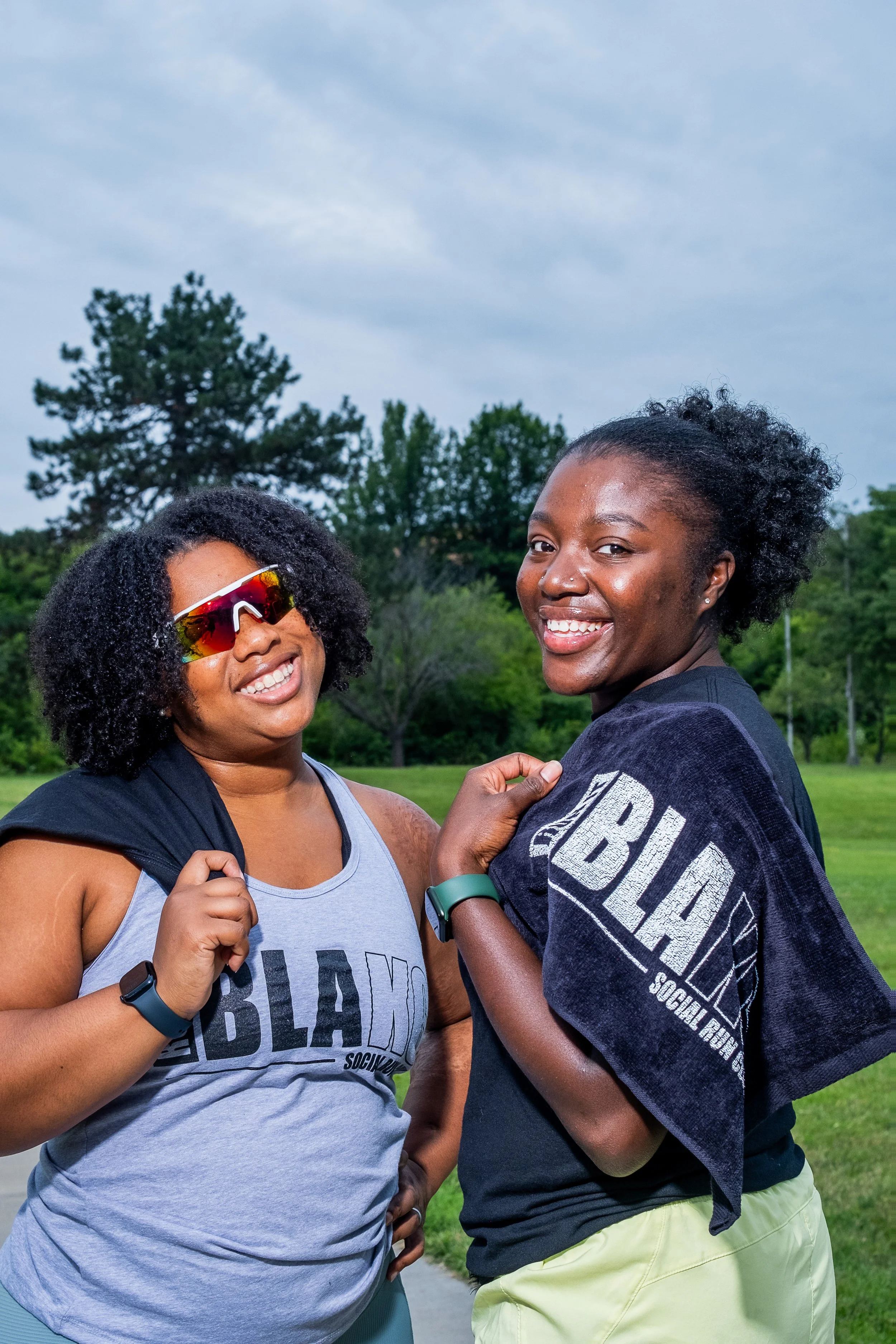 Two women smiling and looking at the camera outdoors in a park, with trees and a cloudy sky in the background. One woman is wearing sunglasses, a gray tank top, and a fitness tracker, and the other is wearing a dark T-shirt with text and a wristwatch