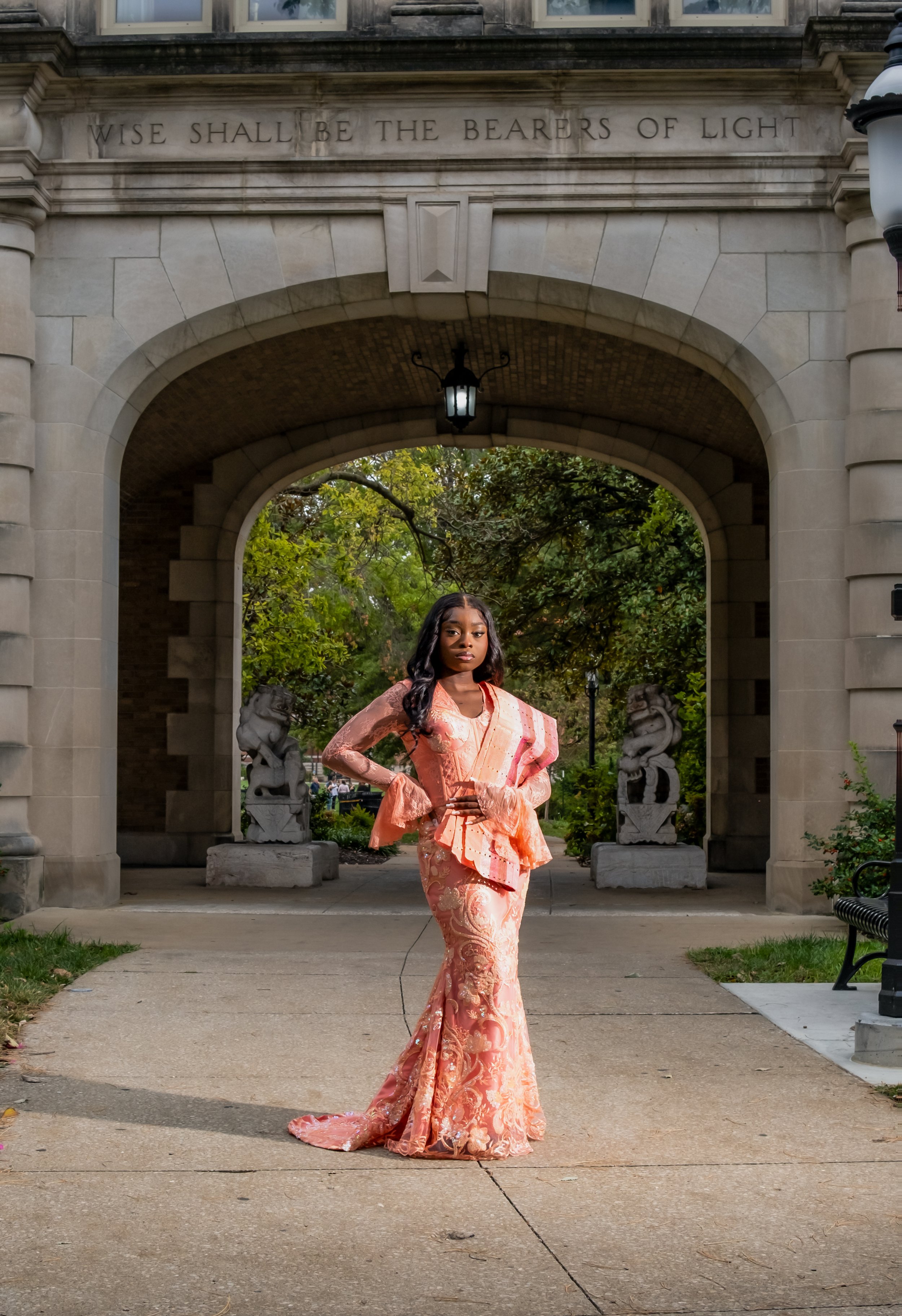 A woman in a pink traditional dress standing under an archway with a quote on it. The quote reads "Wise shall be the bearers of light." There are trees and park features visible in the background.