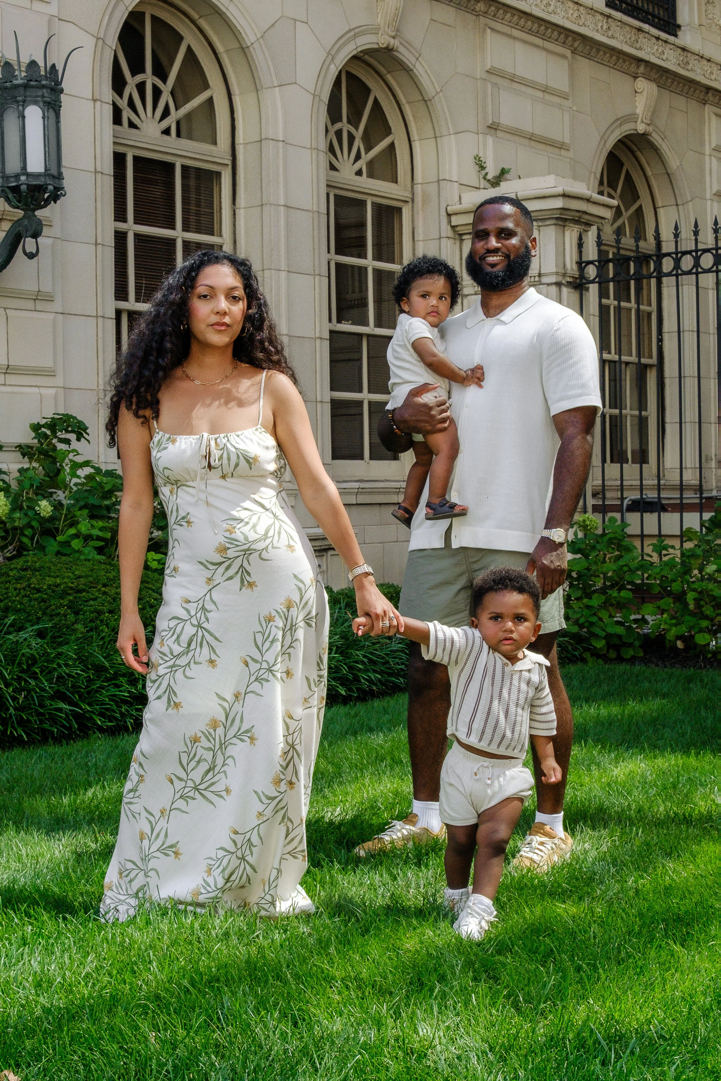 A diverse family of five, including a woman, a man, and three young children, standing on a well-manicured lawn in front of an elegant building with arched windows, ornate stone detailing, and a black wrought-iron fence, enjoying a sunny day.