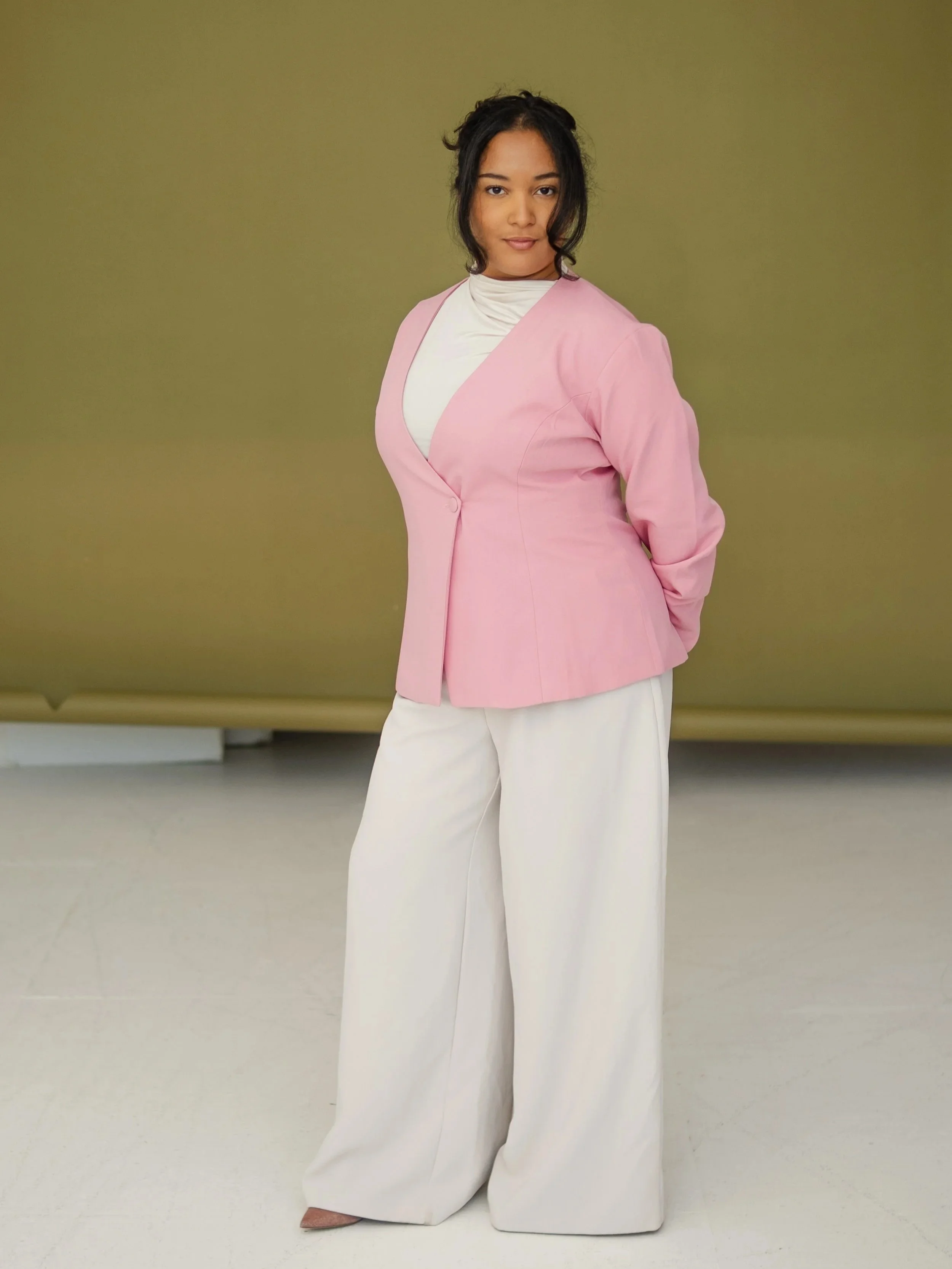 Woman with dark hair in a loose bun, wearing a light pink blazer over a white turtleneck, standing against an olive green background.