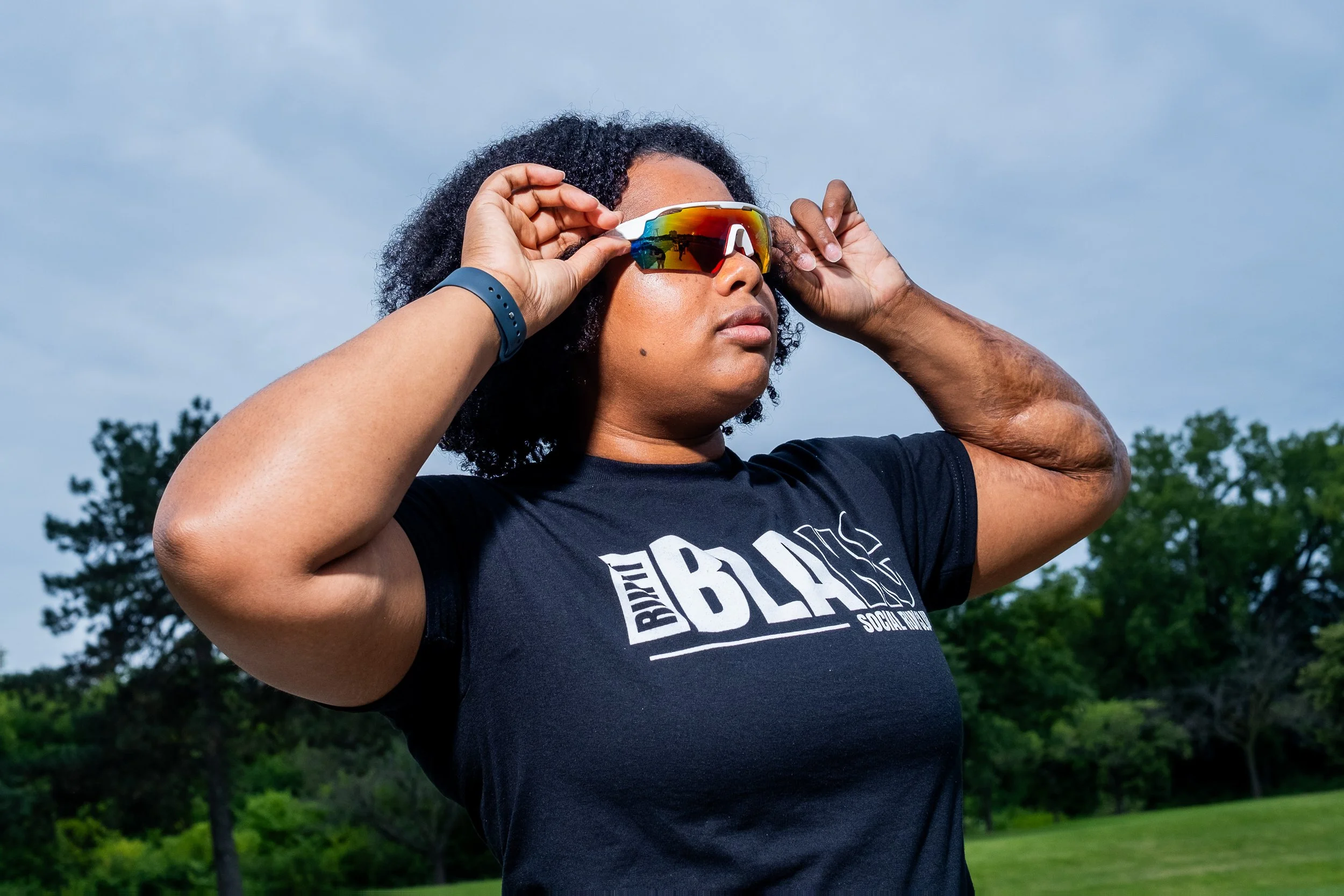A woman with curly hair wearing sunglasses and a black T-shirt with white text, standing outdoors with trees and a cloudy sky in the background, adjusting her sunglasses.