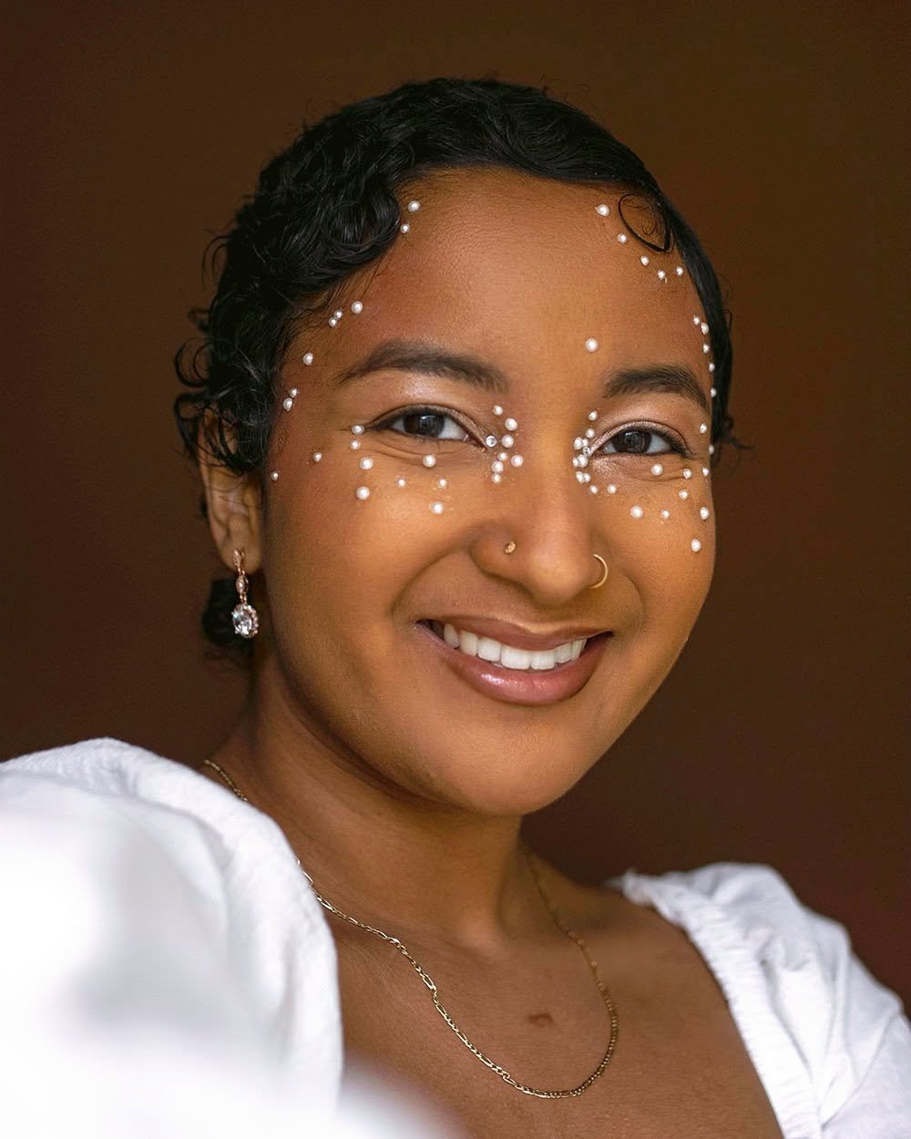 A smiling woman with dark, curly hair adorned with small pearls, wearing pearl earrings and a gold necklace, with white face paint and pearls on her face, set against a dark brown background.