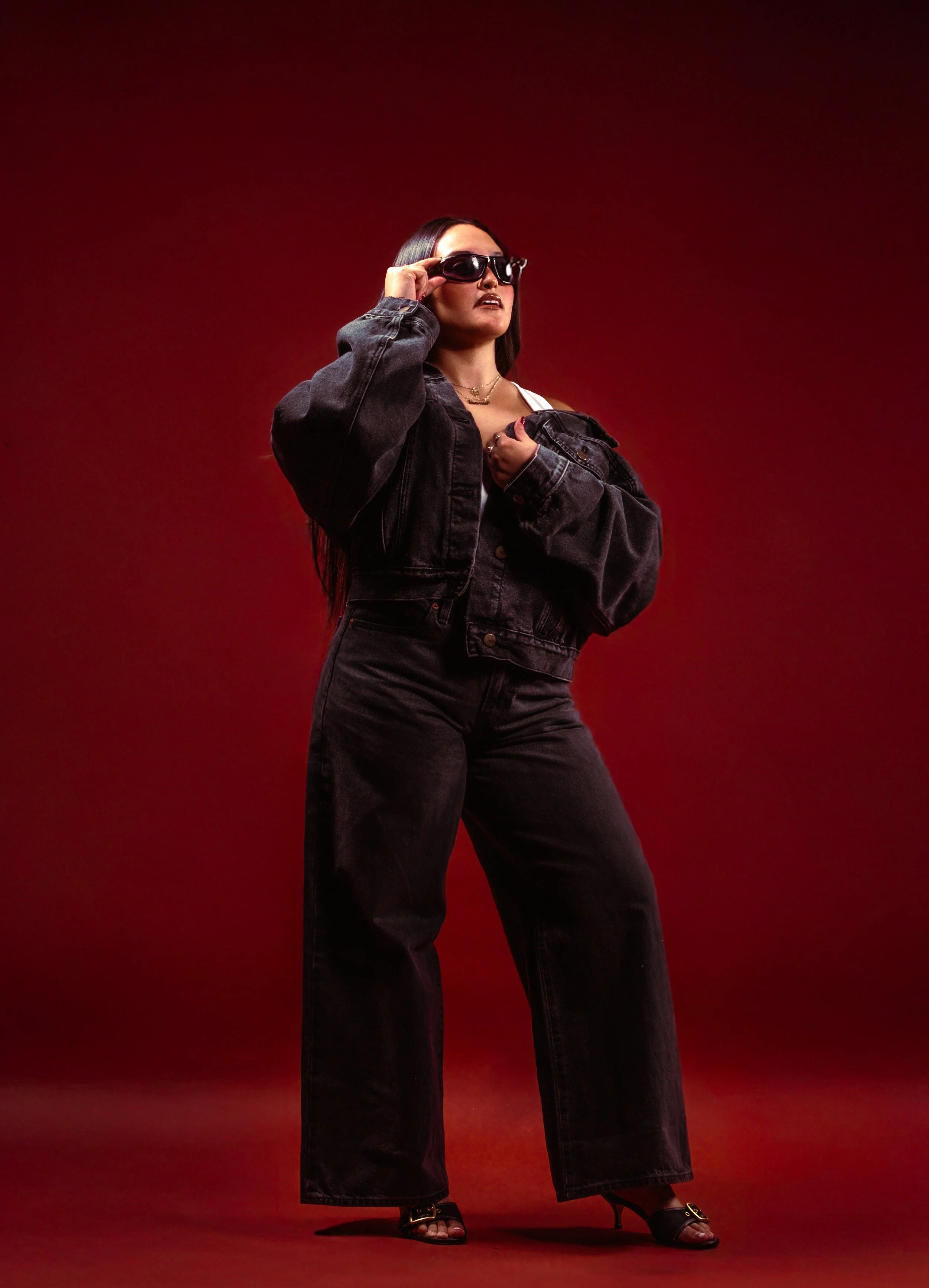 A woman with long black hair wearing sunglasses, a black denim jacket, and black high-heeled shoes stands against a red gradient background.