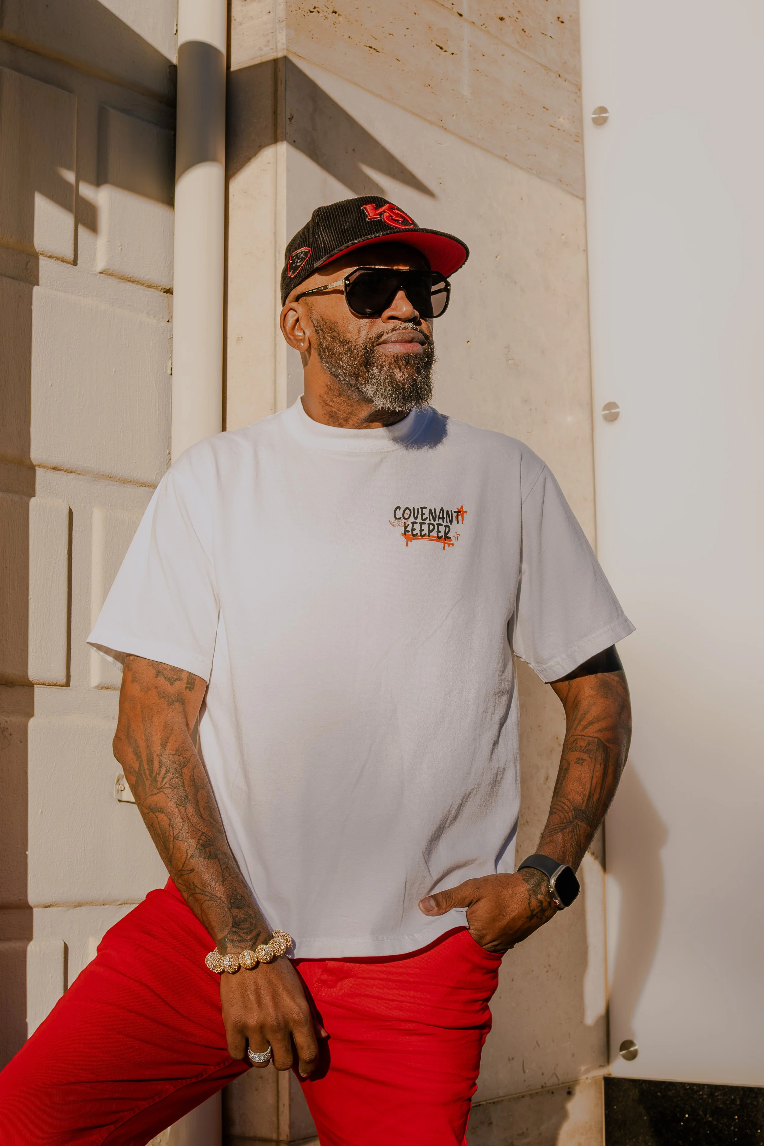 A man wearing a black cap with red accents, black sunglasses, a white T-shirt with black and orange text, red pants, and a gold bracelet, standing against a stone wall with shadows cast by the sunlight.