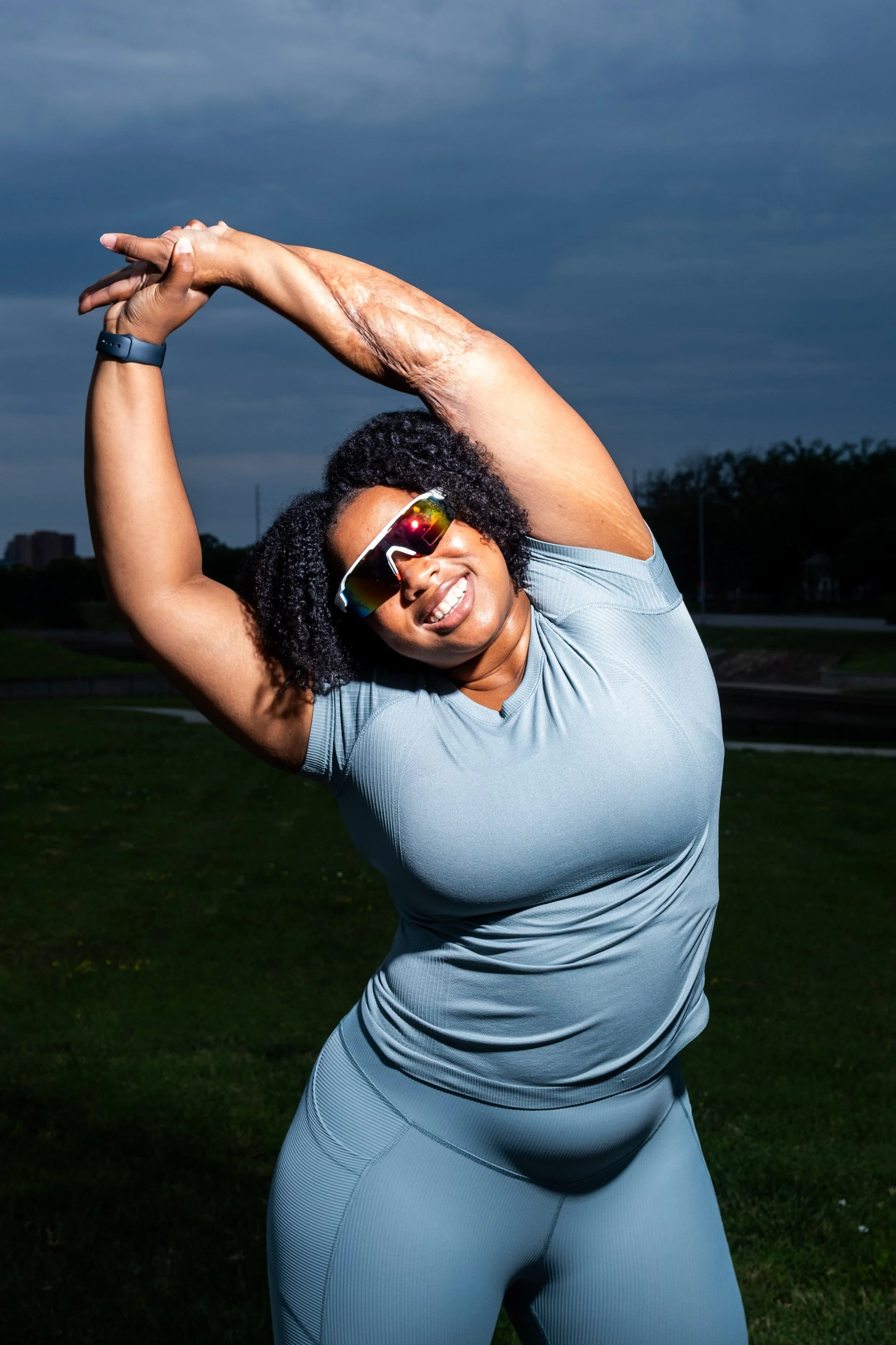 A woman outdoors stretching, wearing sunglasses and a light gray workout outfit, with a cloudy sky in the background.
