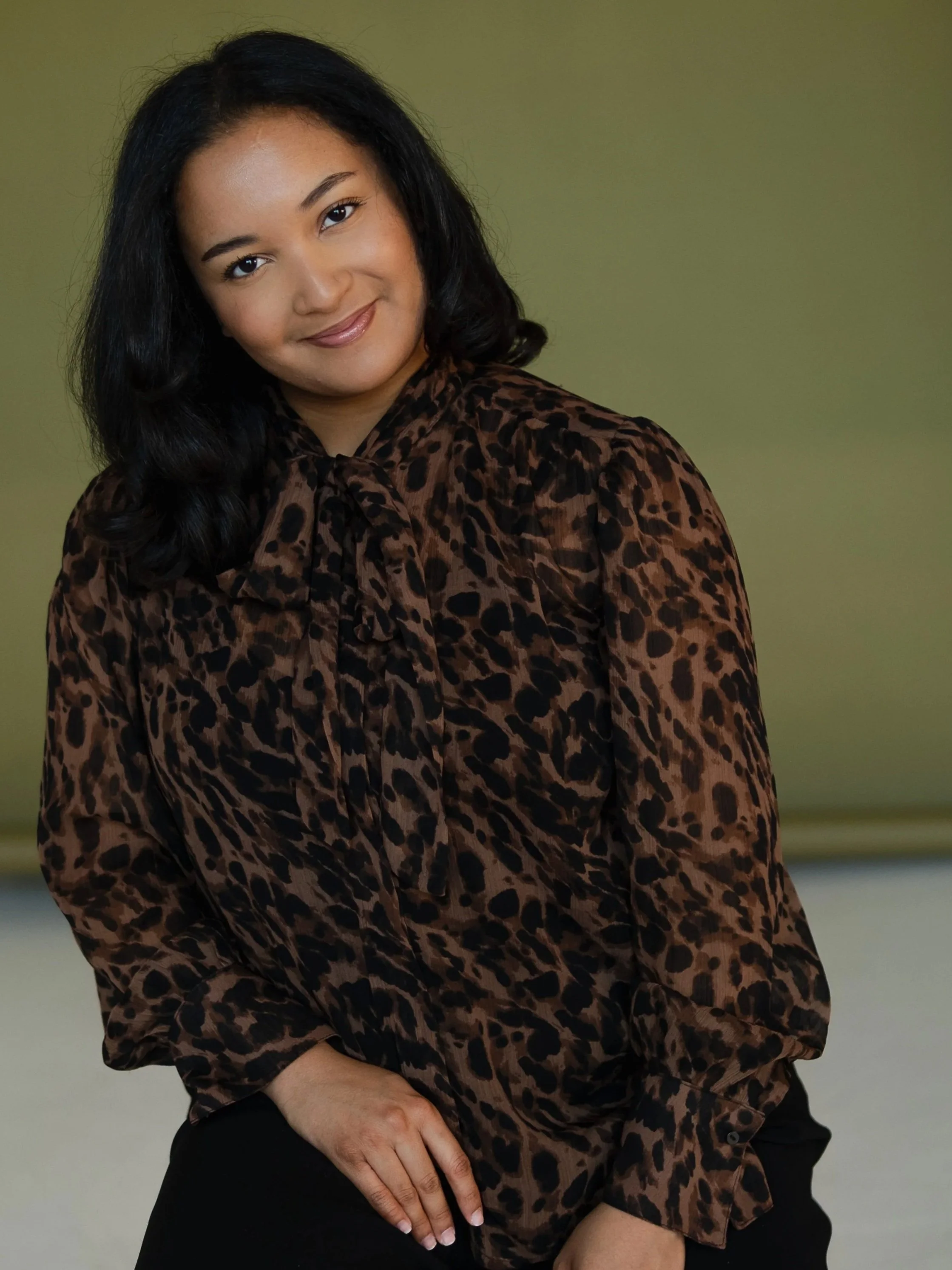 A woman with black hair wearing a leopard print blouse, sitting with her left hand resting on her lap against a blurred background.