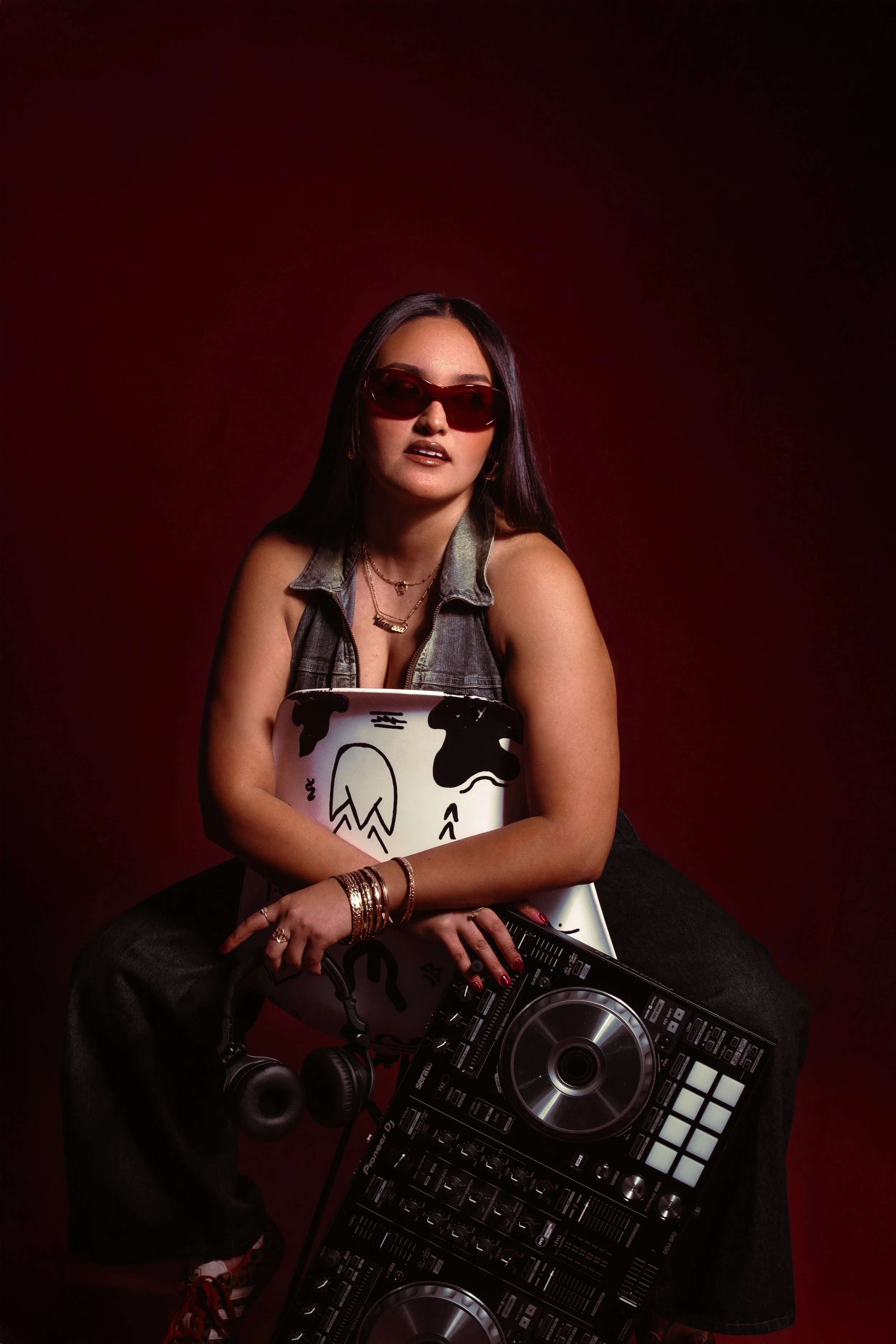 A woman with long dark hair wearing sunglasses and a denim vest, holding a laptop with a cow pattern and a DJ controller, seated against a dark red background.