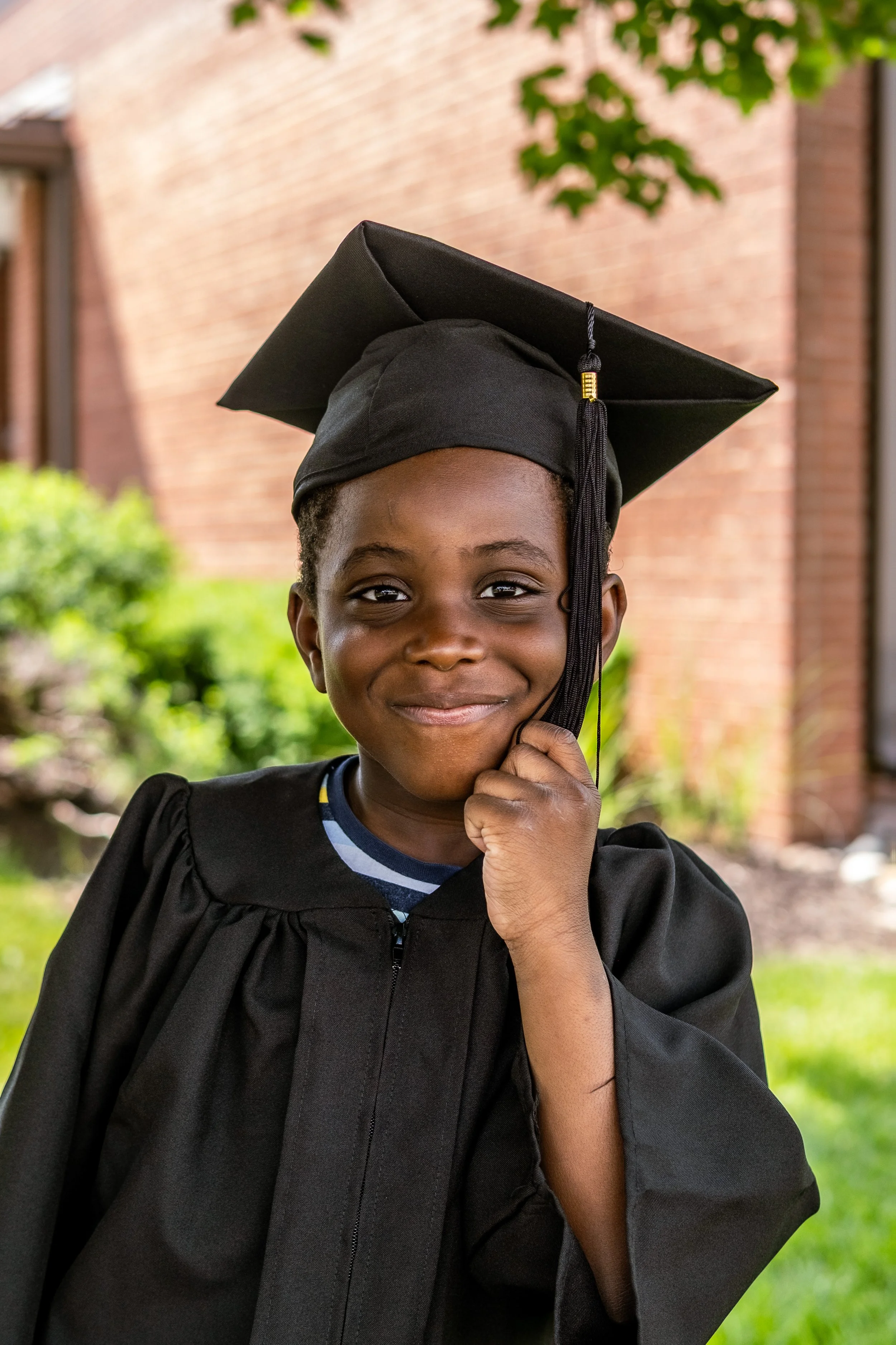 A young girl dressed in a black graduation gown and cap, holding the tassel, standing outdoors in front of a brick building and greenery, smiling at the camera.