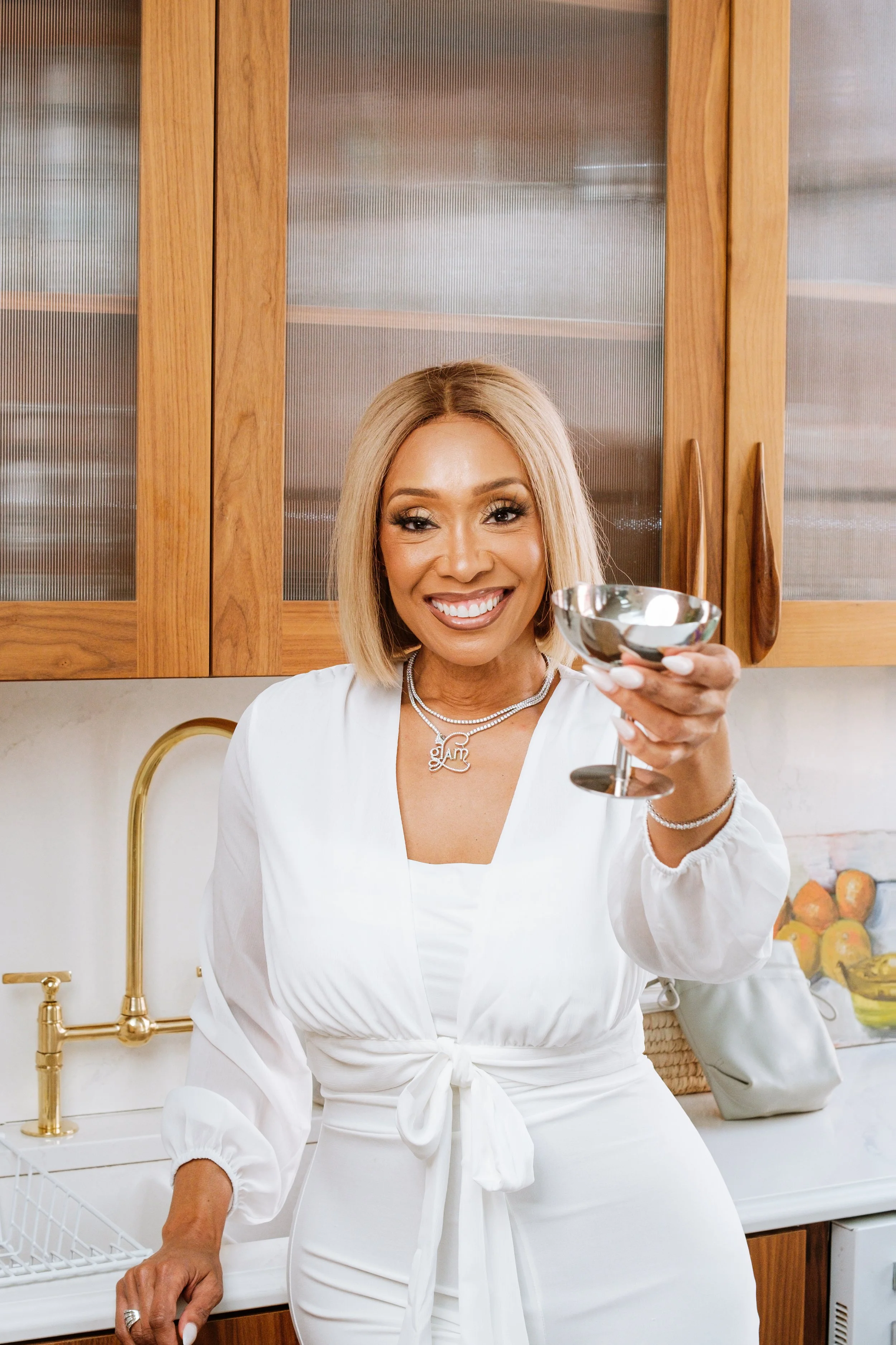 A smiling woman with blonde hair holding a silver cocktail shaker in a kitchen with wooden cabinets and gold fixtures.