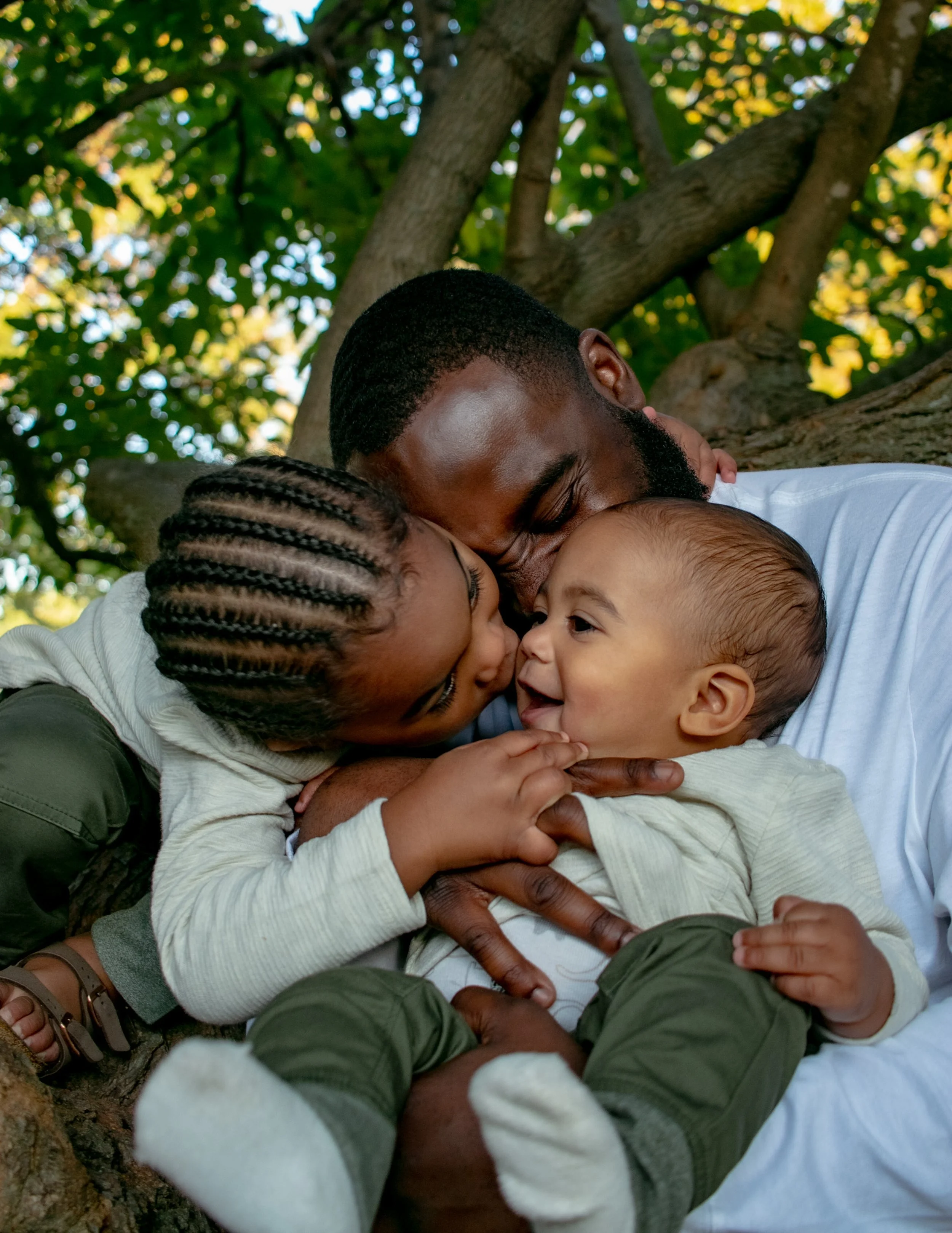 A man and two children hugging each other under a tree, with the children smiling and the man resting his head on the child's head.