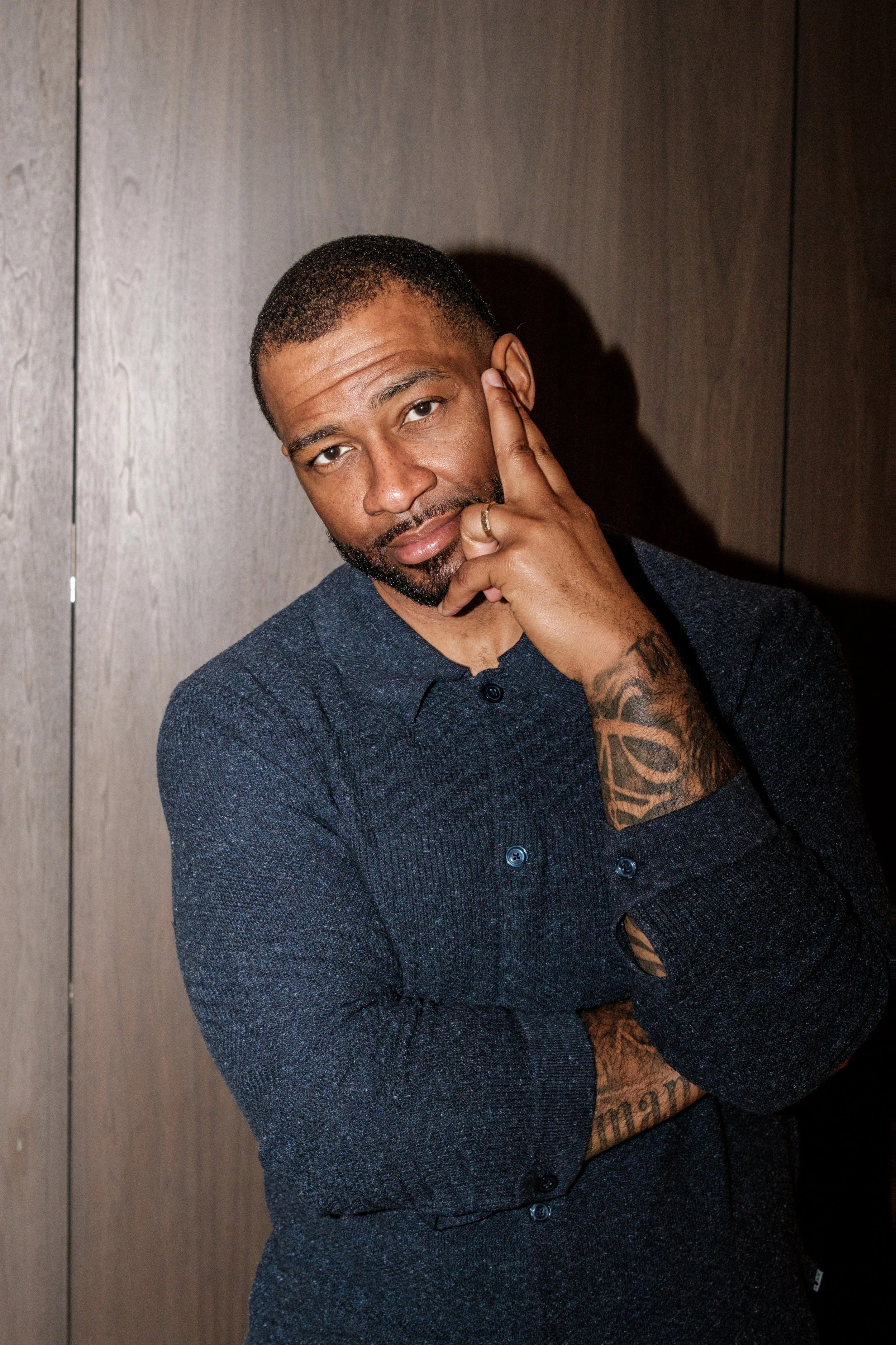 A man with short hair, beard, and tattoos on his arm, wearing a dark blue sweater, is posing with his finger on his face near his eye, against a wooden background.