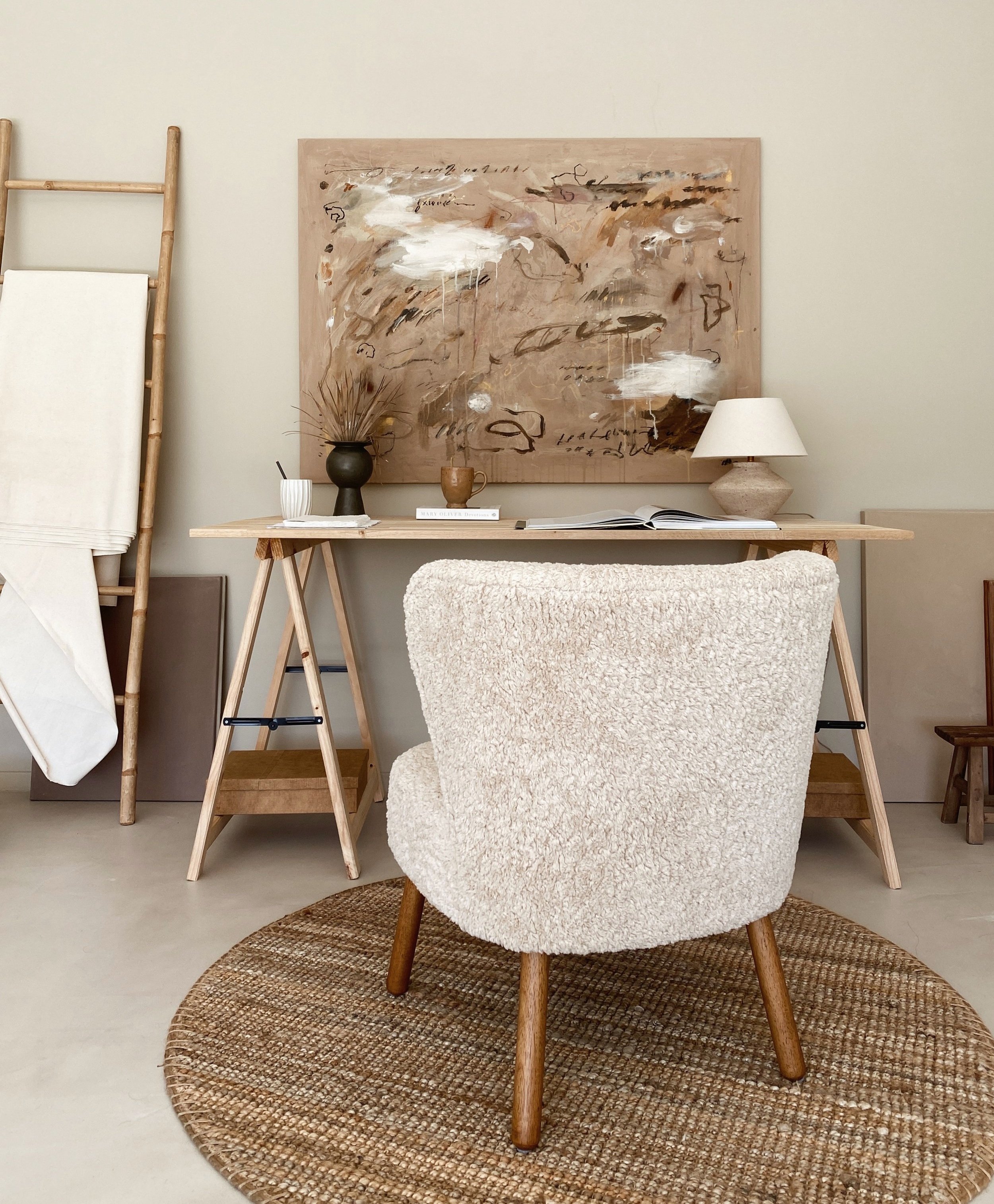 A cozy home office with a white plush armchair on a round woven rug facing a simple wooden desk. The desk has a table lamp with a white shade, a small potted plant, a coffee mug, and open books. Behind the desk, there's a large abstract painting with