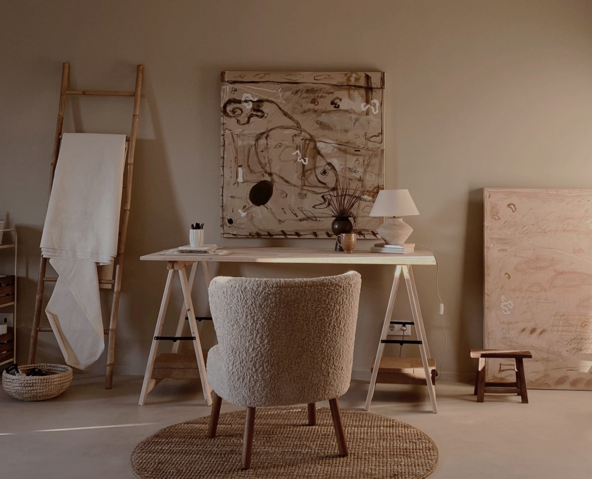 A cozy home office with a light wood desk, a plush chair, trendy textile rugs, and modern decor including artwork, a table lamp, and decorative vases.