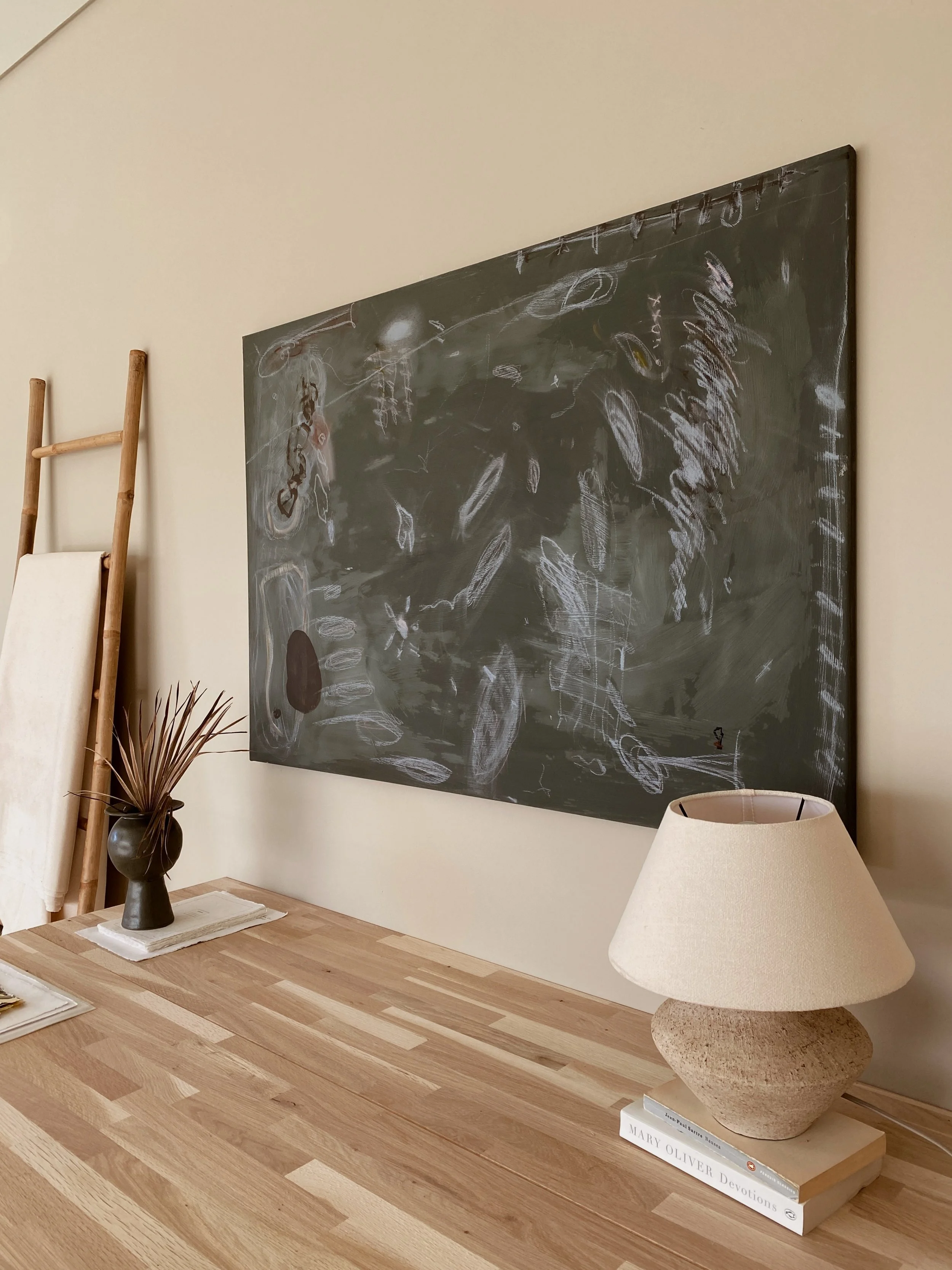A wooden desk with a lamp, books, and a vase with dried foliage, against a beige wall with a black chalkboard.