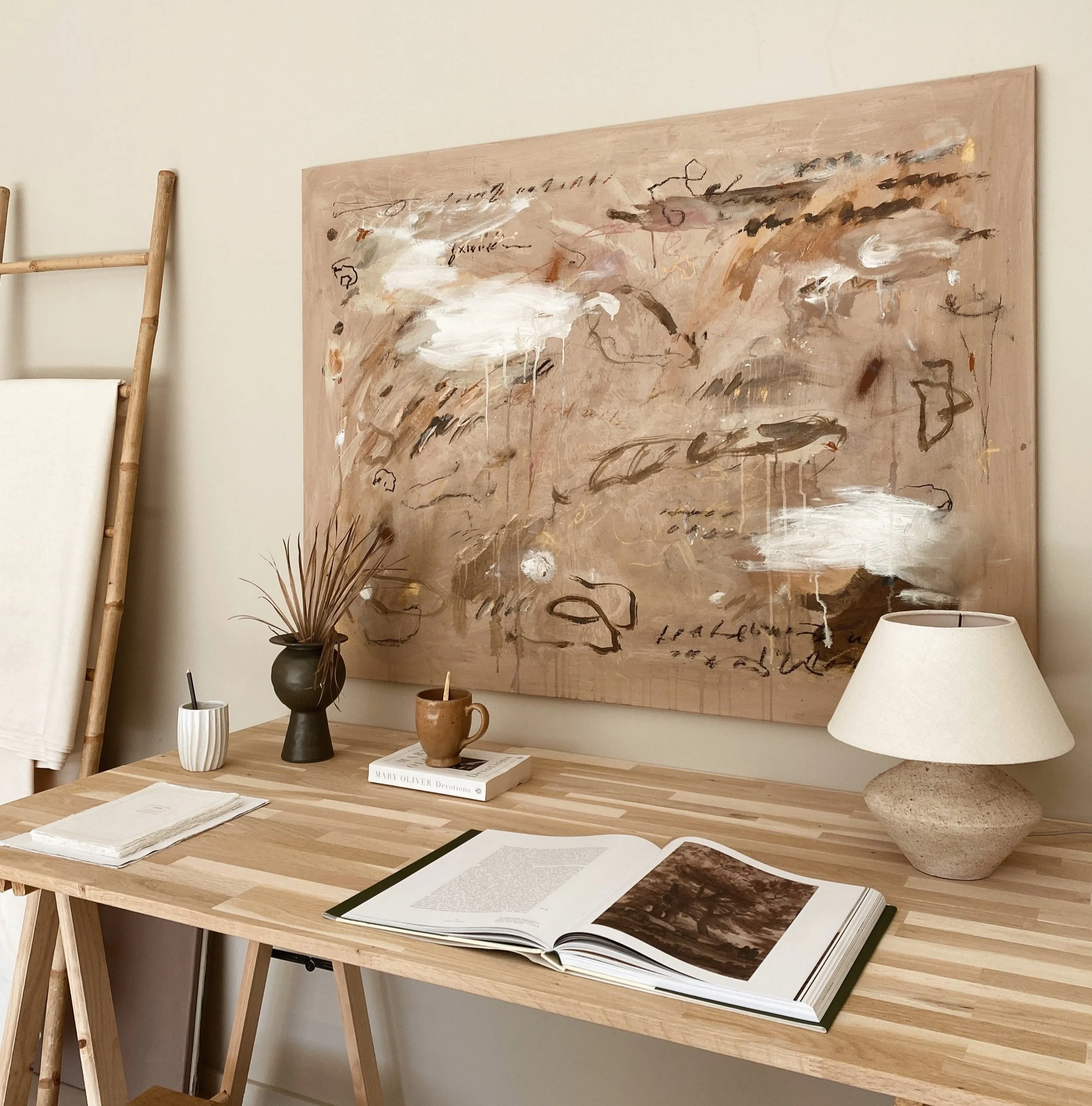 A wooden desk with books, a cup, and a table lamp, against a wall with an abstract painting.