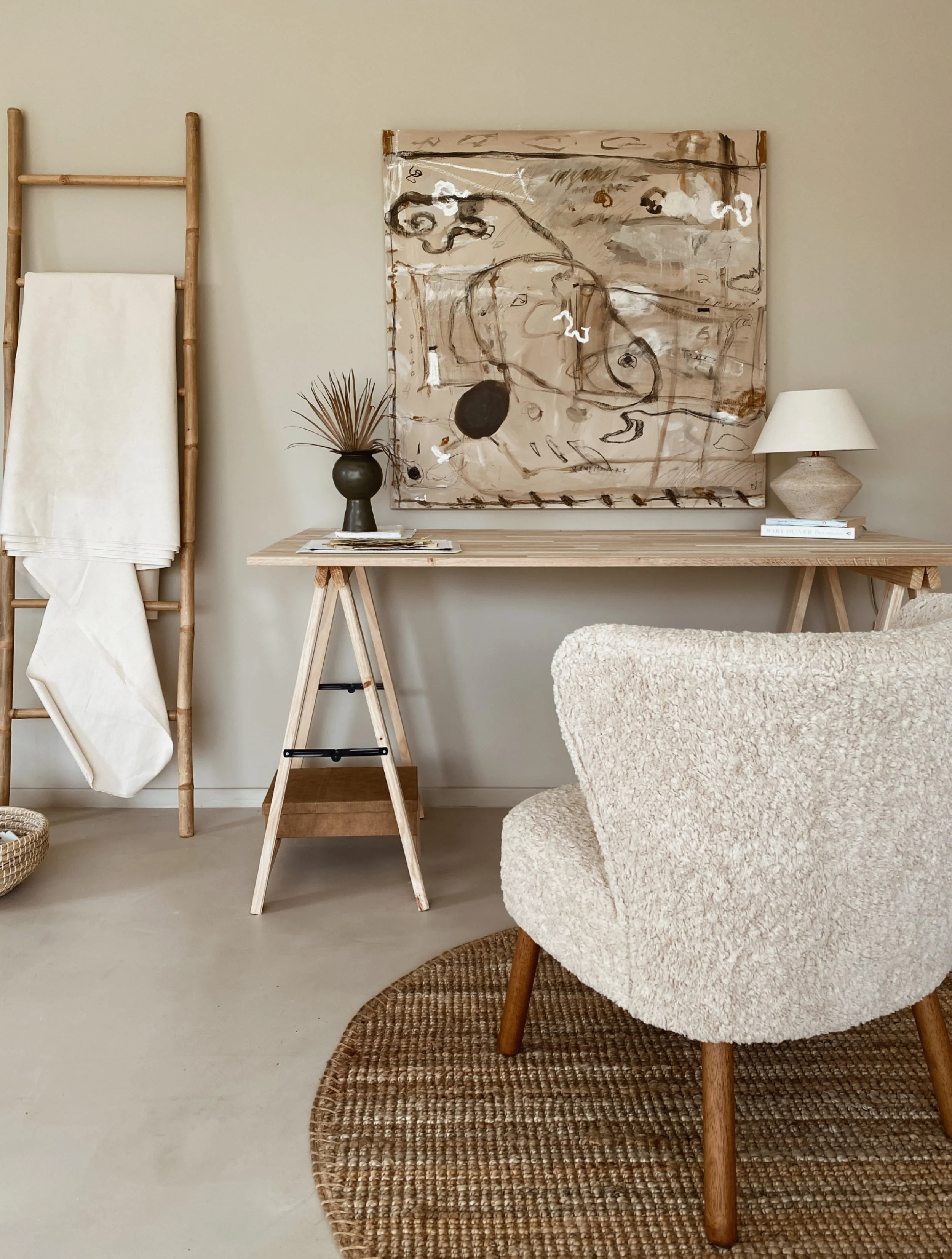 Interior of a room featuring a wooden ladder with a white cloth, a wooden table with a black vase and a stack of magazines, a beige armchair on a round woven rug, a large abstract painting on the wall, a white table lamp, and a small stack of white b