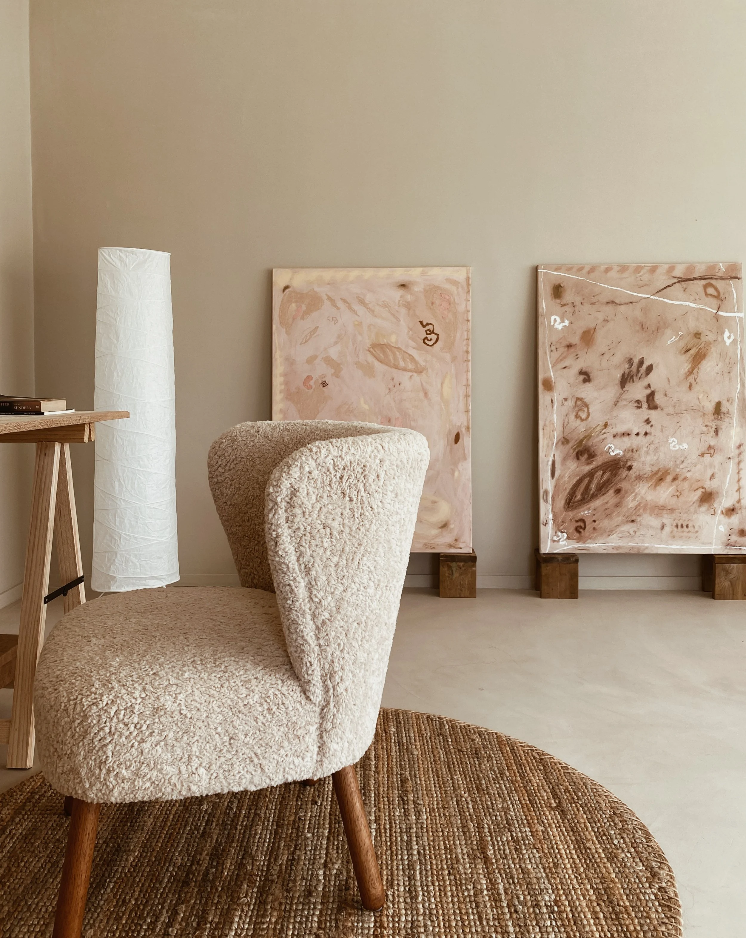 Interior room with a plush beige armchair, side table, tall white lamp, and two pink abstract paintings on wooden stands.