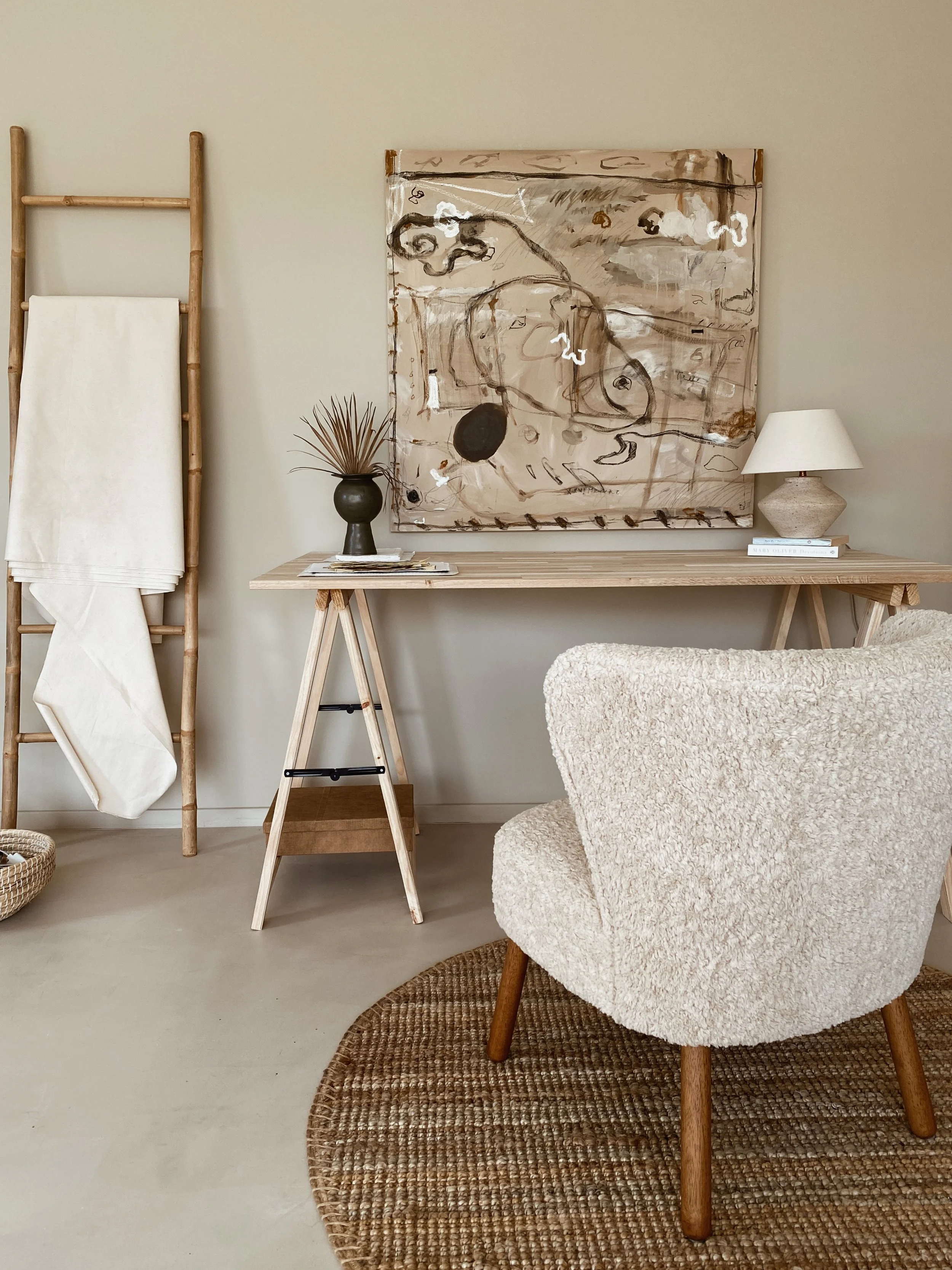 Interior of a room with a beige armchair, a wooden desk, a patterned rug, a table lamp, a black vase with dried plants, an abstract painting, a wooden towel rack with a white towel, and a woven basket.