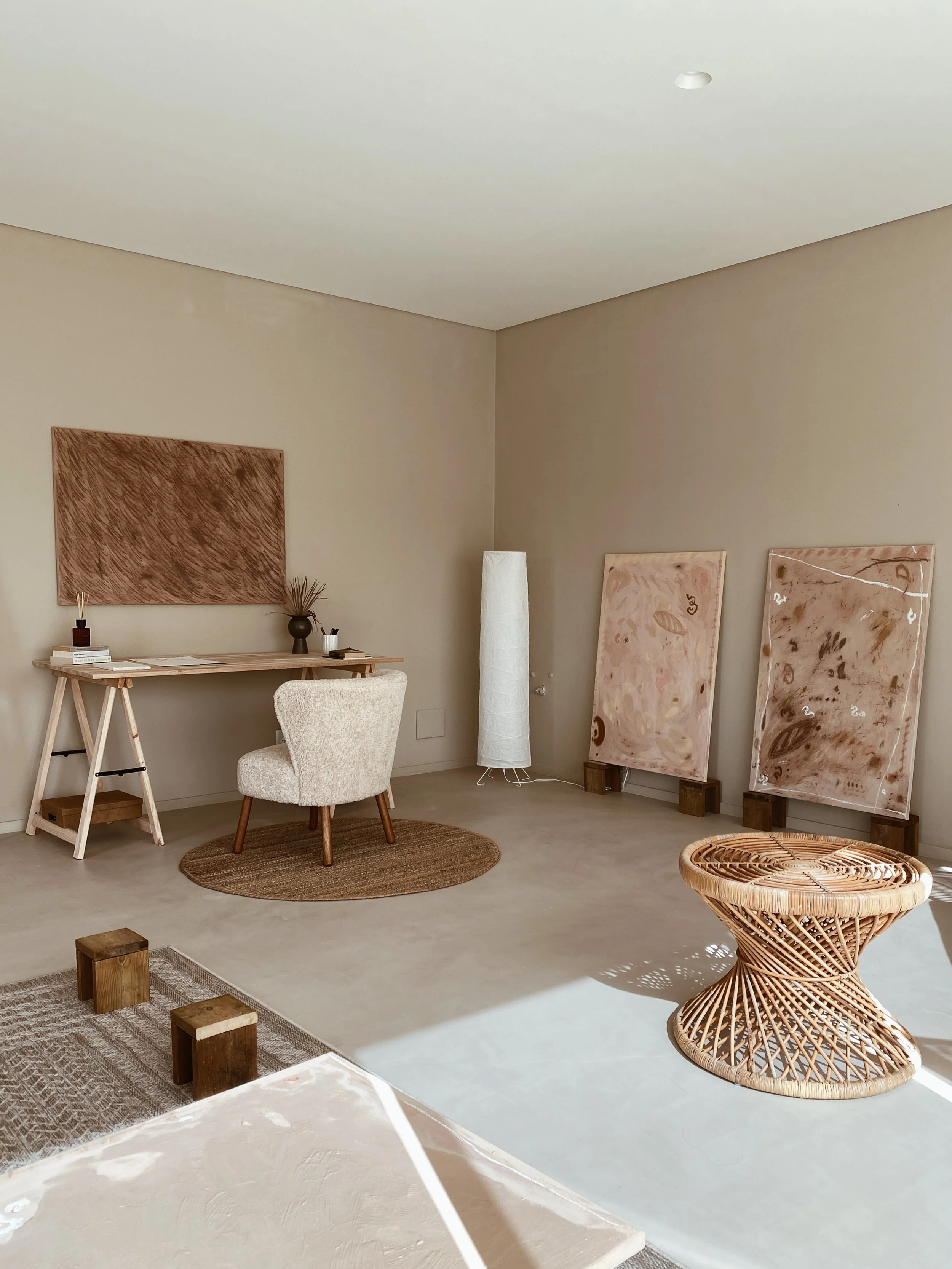 Minimalist interior room with a beige chair on a round rug, two large abstract pink paintings on wooden stands, a rattan stool, a tall white floor lamp, and a small wooden desk with decor, set against beige walls and cream carpet.