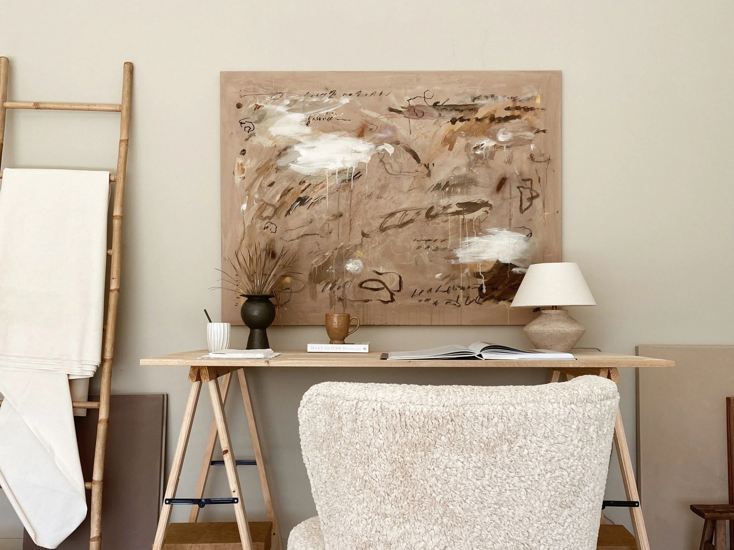 Interior home office with a light wood desk, a cozy cream armchair, an abstract painting on the wall, a table lamp, and decorative items such as a vase with dried plants and a cup.