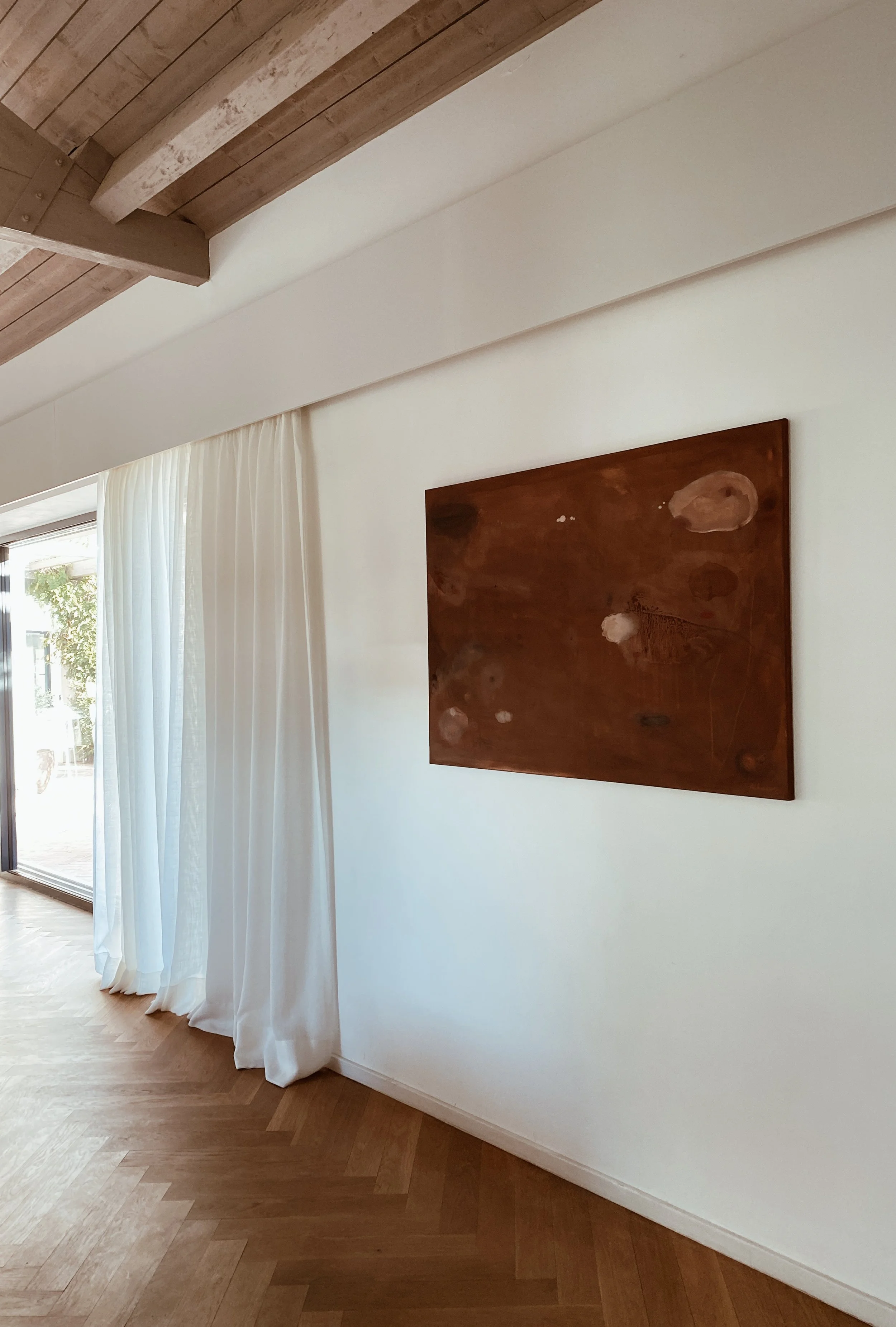 Interior of a room with white walls, wooden ceiling beams, light-colored curtains, and a wooden floor, featuring an abstract brown painting on the wall.