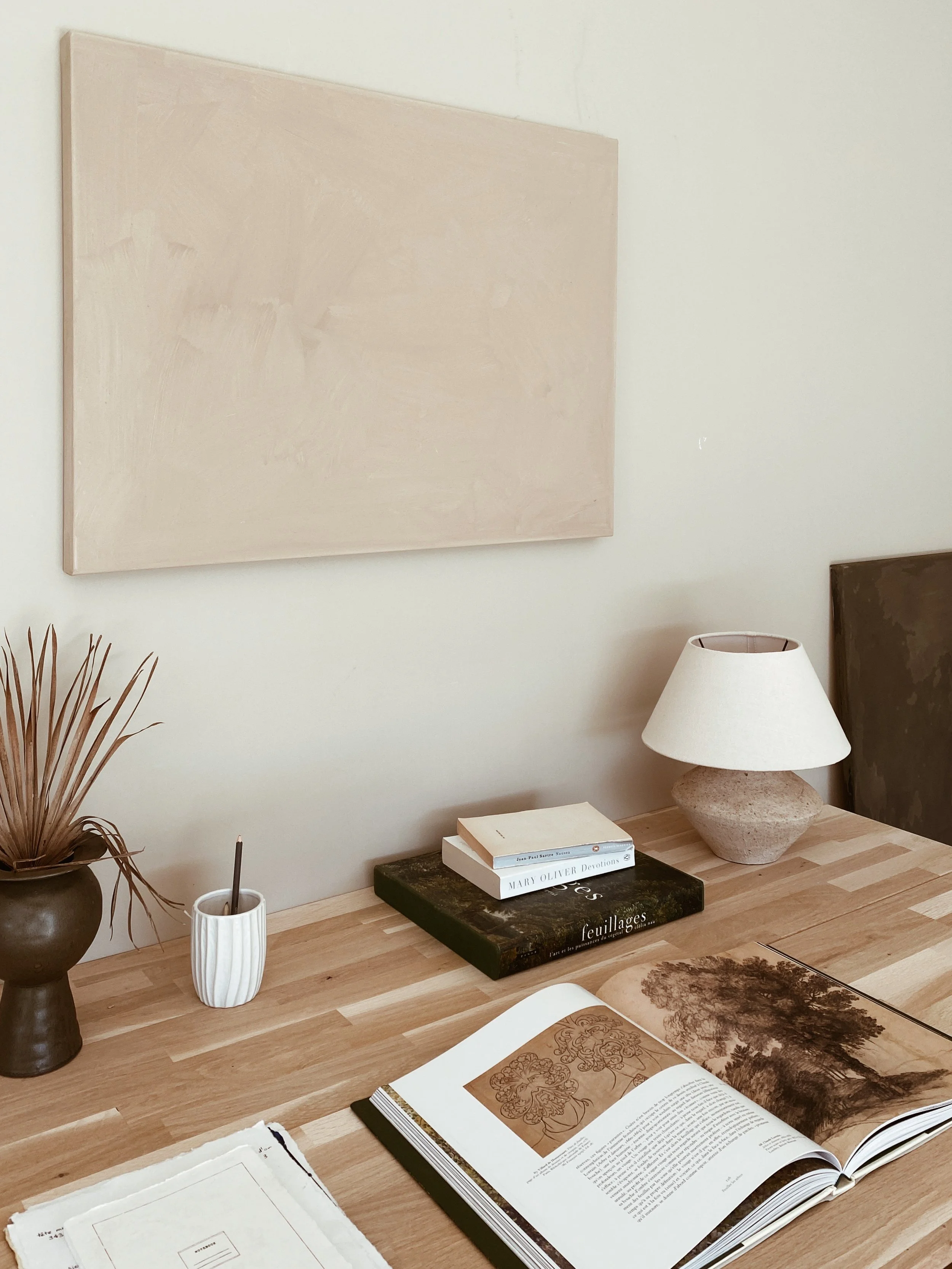 A wooden table with an open art book, small stacked books, a white mug with a pen, a clay vase with dried leaves, and a table lamp with a stone base against a light-colored wall with a blank canvas.
