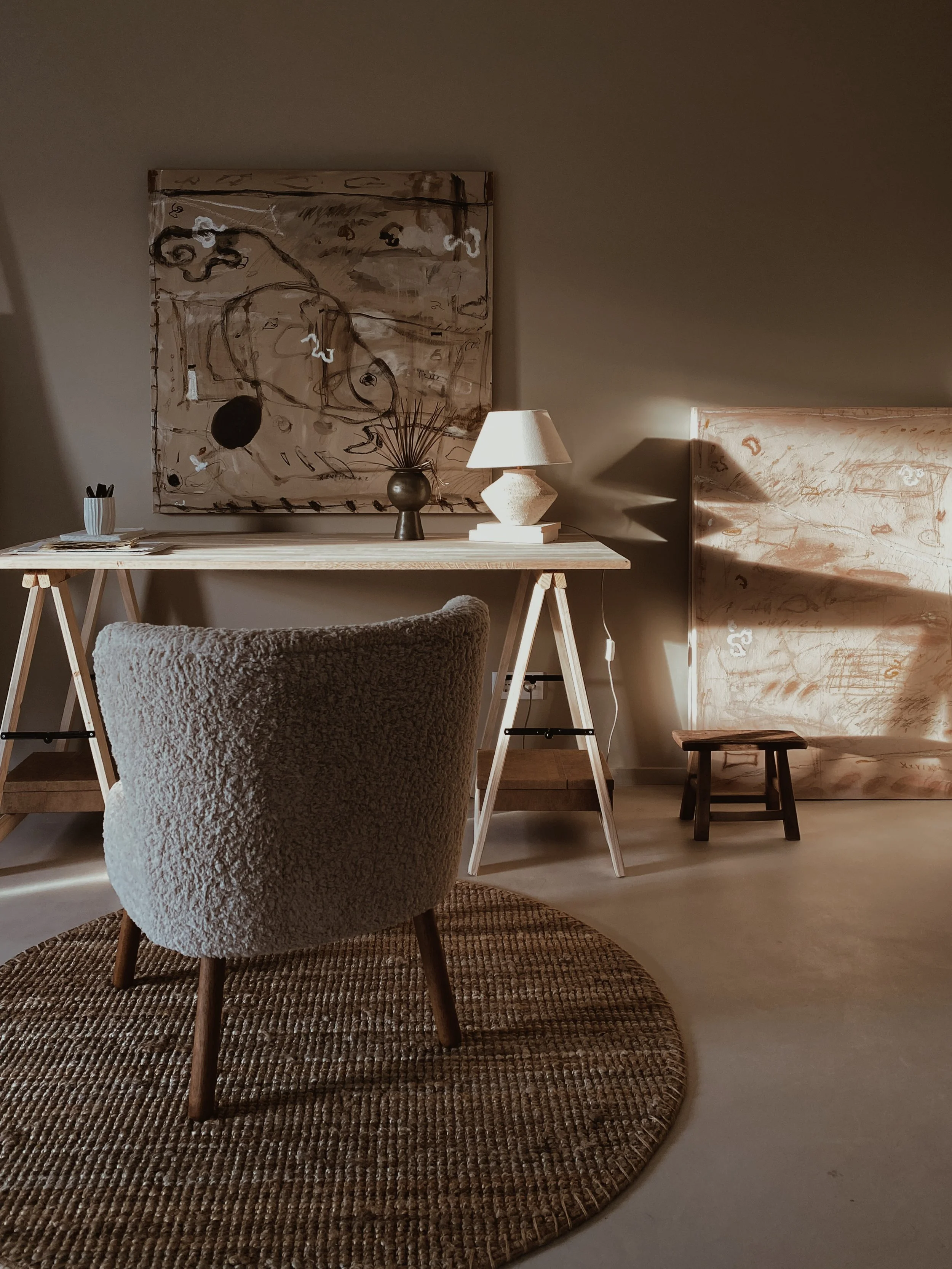 A cozy, minimalist home office with a textured beige armchair on a round woven rug, a wooden desk with a white lampshade, a black vase, and a container with pens. Pastel abstract artwork hangs on the wall behind the desk, and a small wooden stool is 