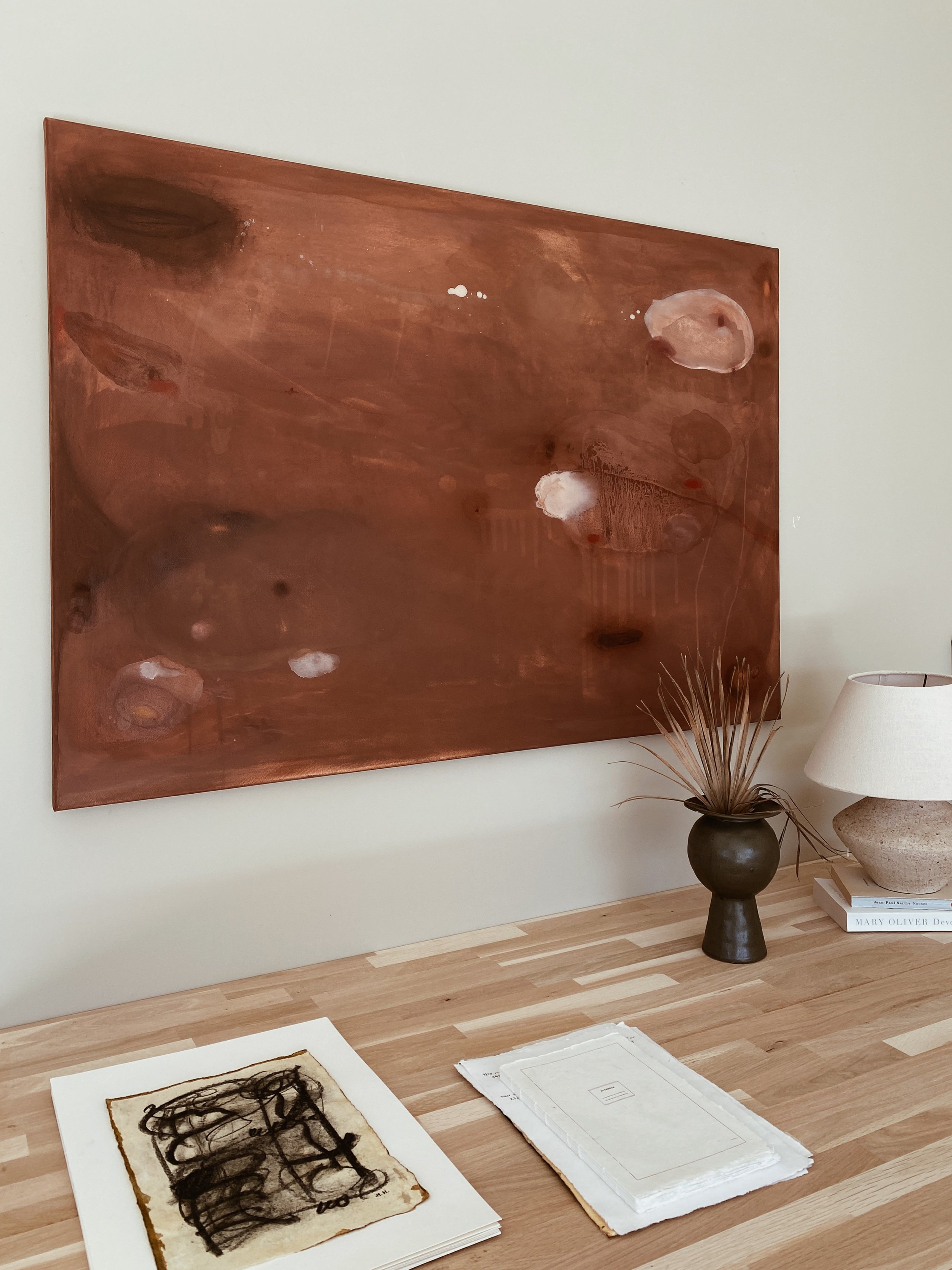 A large abstract painting with a reddish-brown background and various white and darker brown blotches hangs on a white wall above a wooden table. On the table, there is a dark vase with dried plant stems, a table lamp with a textured base, a stack of