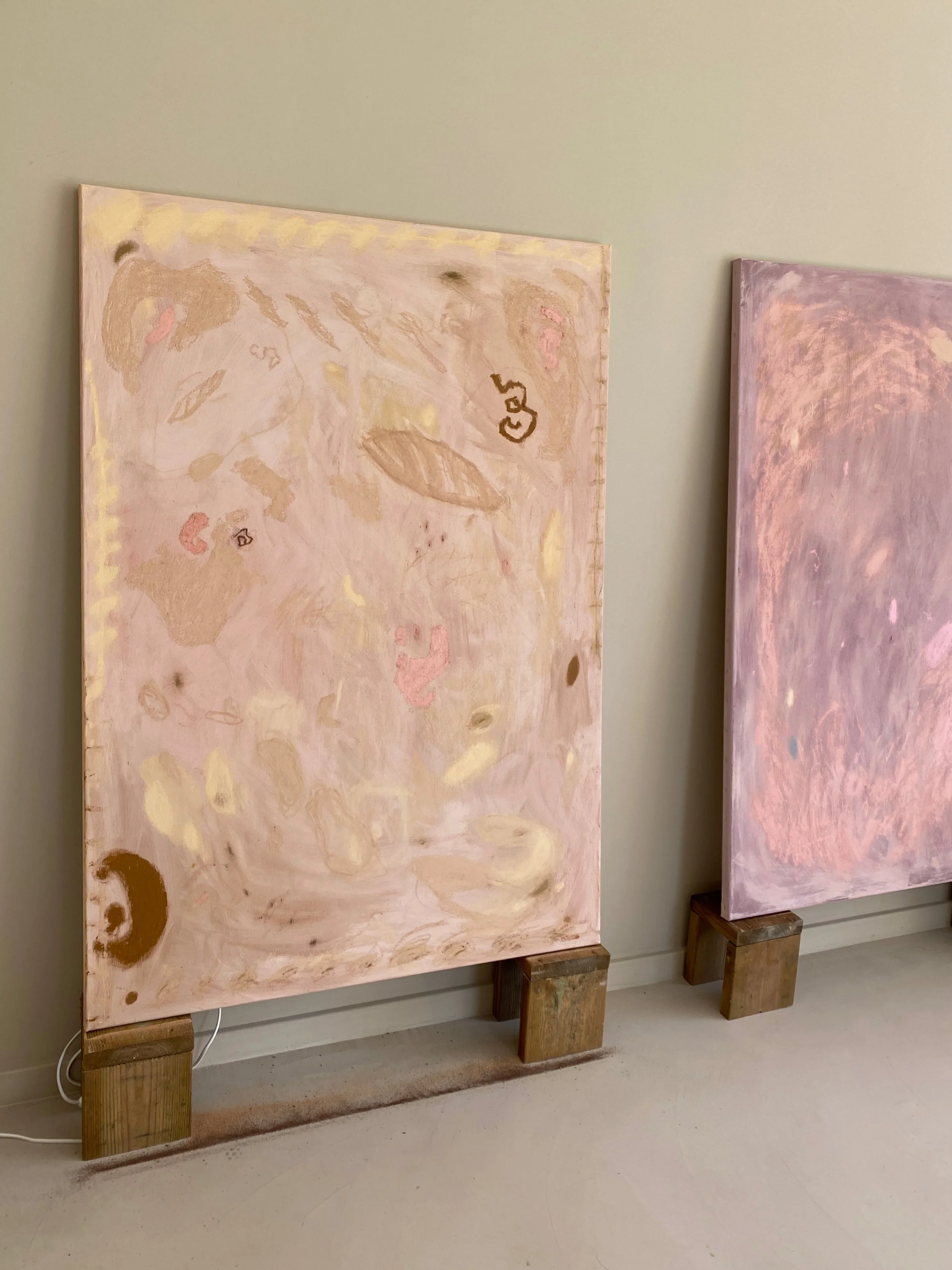 Two large abstract paintings leaning against a wall on wooden blocks, featuring soft pink and beige tones with organic shapes.