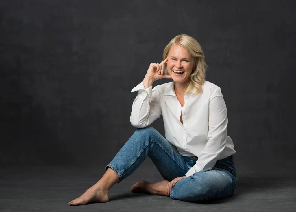 Tanja Törnkvist istuu lattialla valkoisessa paidassa ja farkuissa nauraen, tumma studiotausta