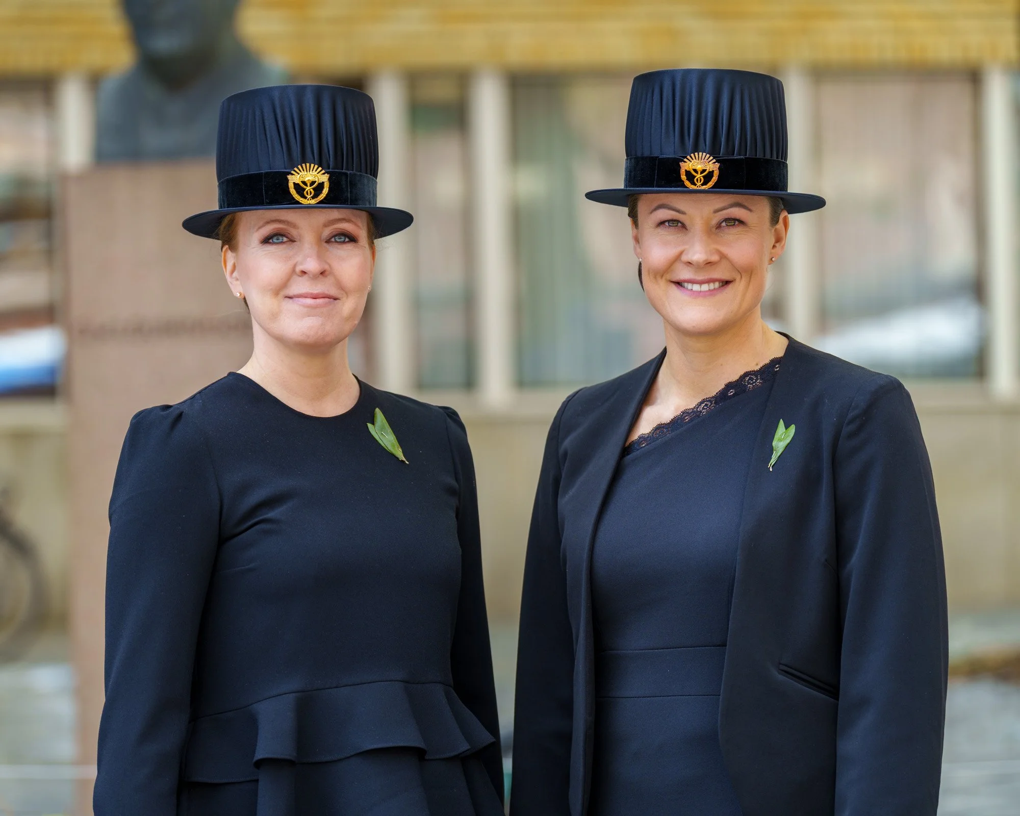 Kaksi naista pukeutuneina mustiin juhlapukuihin, päähineet päähissään, ja houkuttelevat hymyt, kaupungin rakennusten taustalla.