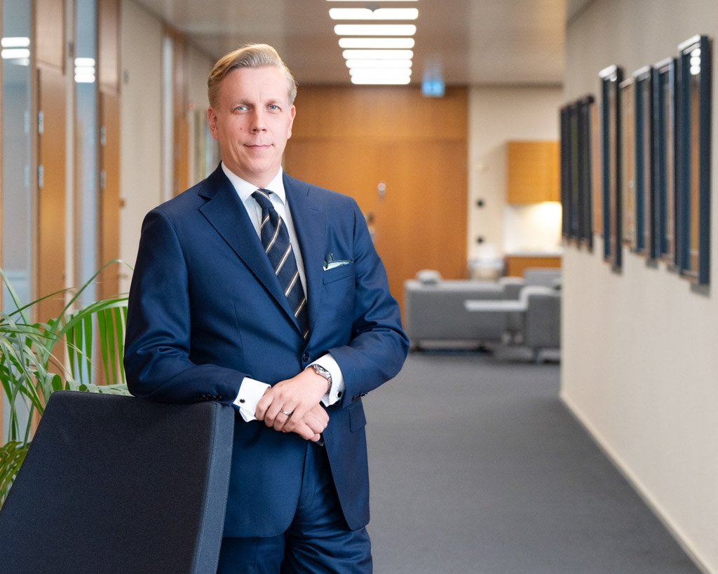 Mies sinisessä puvussa toimiston käytävällä nojaa tuolin selkänojaan