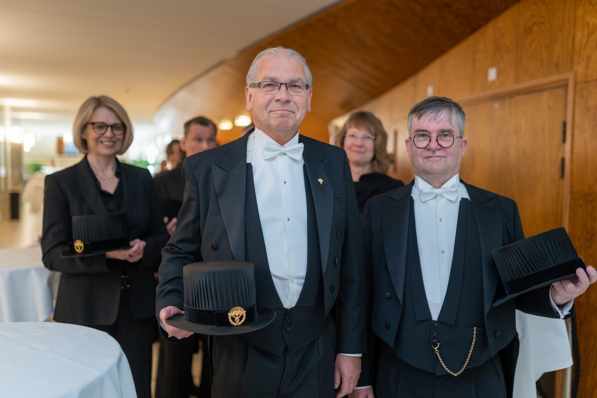 Ihmiset maalaissaleissa ja juhlapuvuissa tapahtumassa, kaksi miestä etualalla pitää päähattuja ja hymyilee, taustalla naisia ja mies nauravat. Tausta on puisia seiniä ja ikkunatettu.