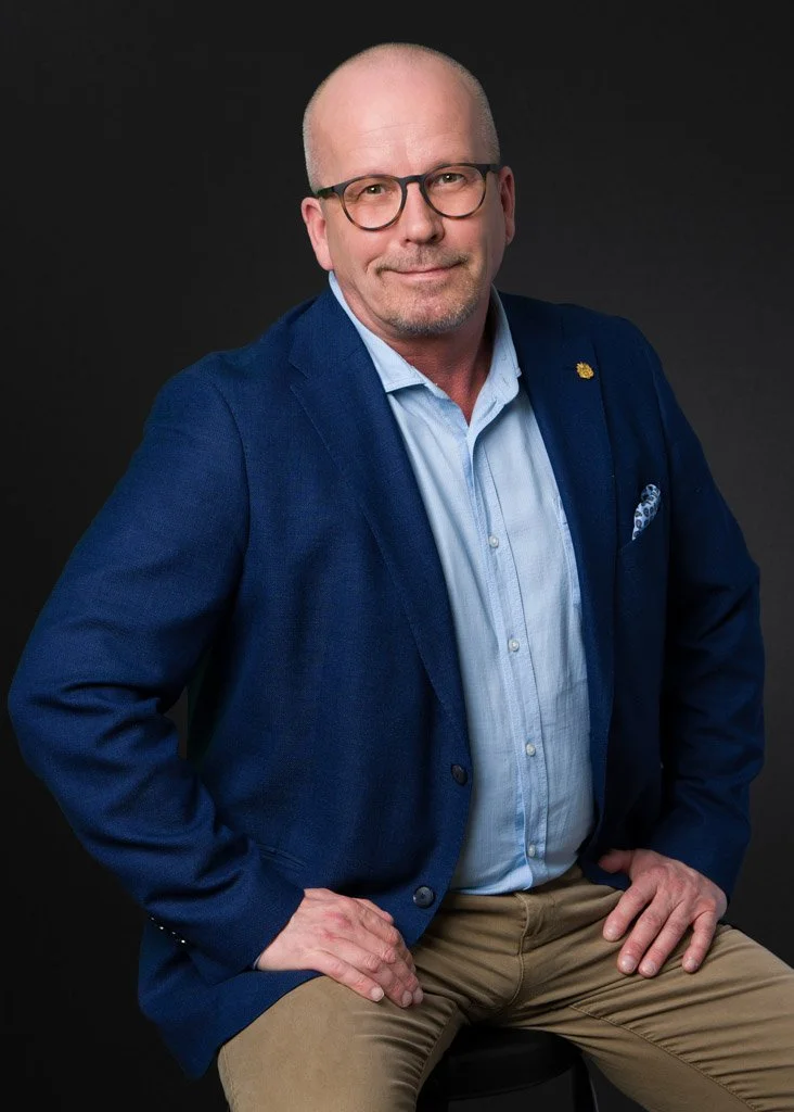Mies sinisessä bleiserissä istuu jakkaralla, tumma tausta