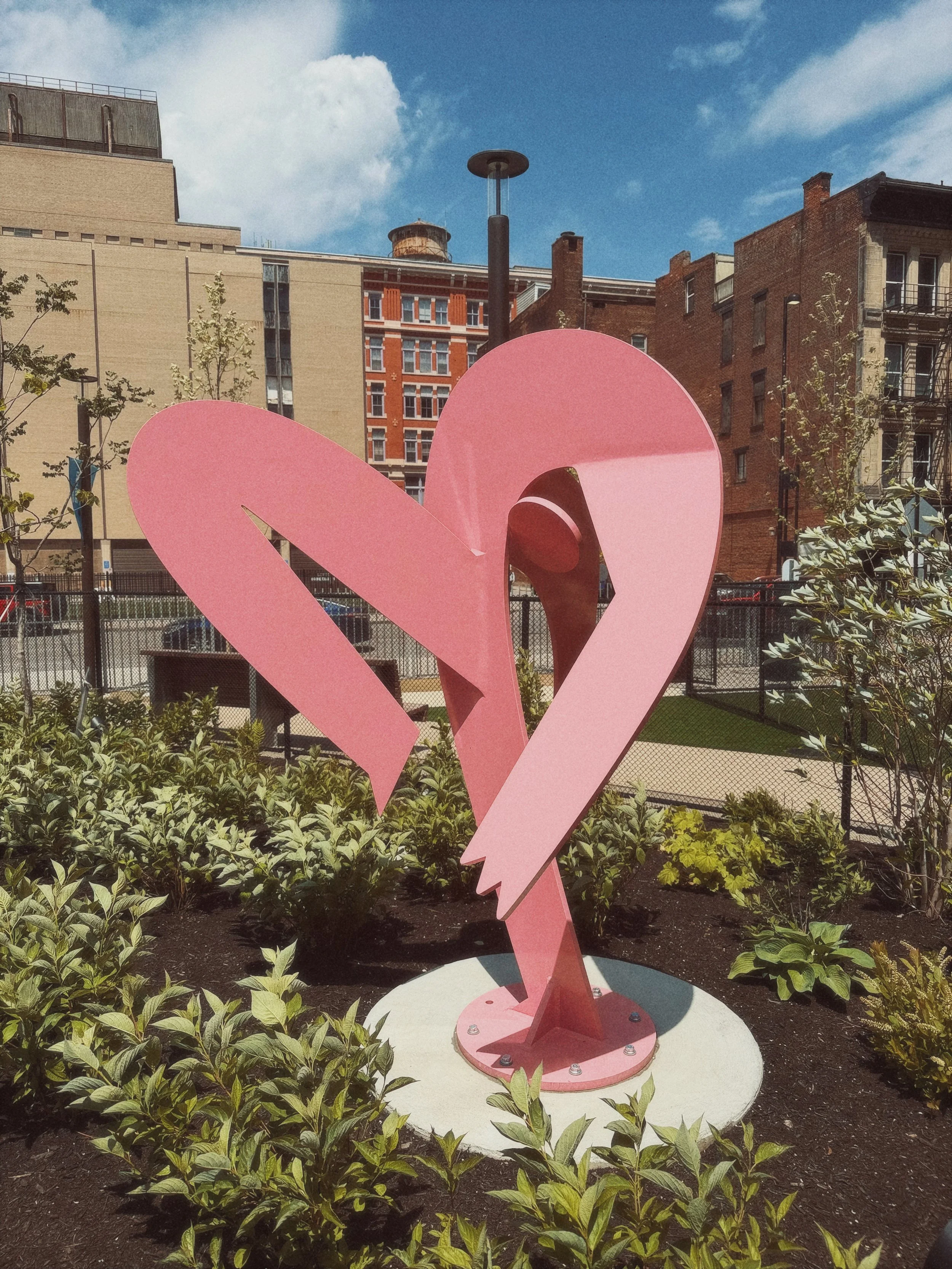 A pink sculpture of a flamingo in a city park with buildings, trees, and a blue sky in the background.