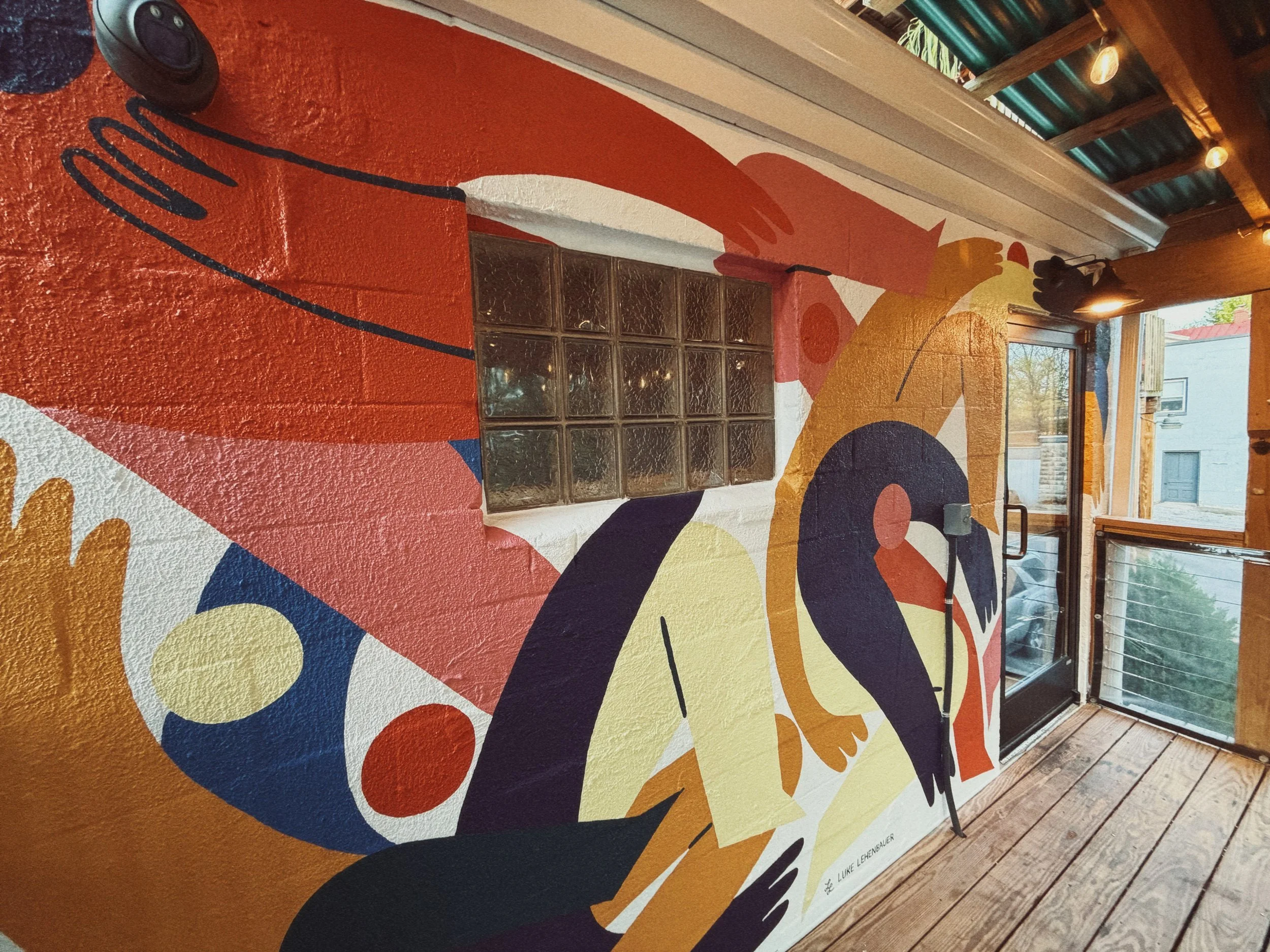 Colorful abstract mural painted on a wall with various shapes and patterns including circles, curves, and lines in bold colors like red, yellow, blue, and black. The mural is situated next to large glass block windows and a sliding glass door, with a wooden ceiling and small ceiling lights.