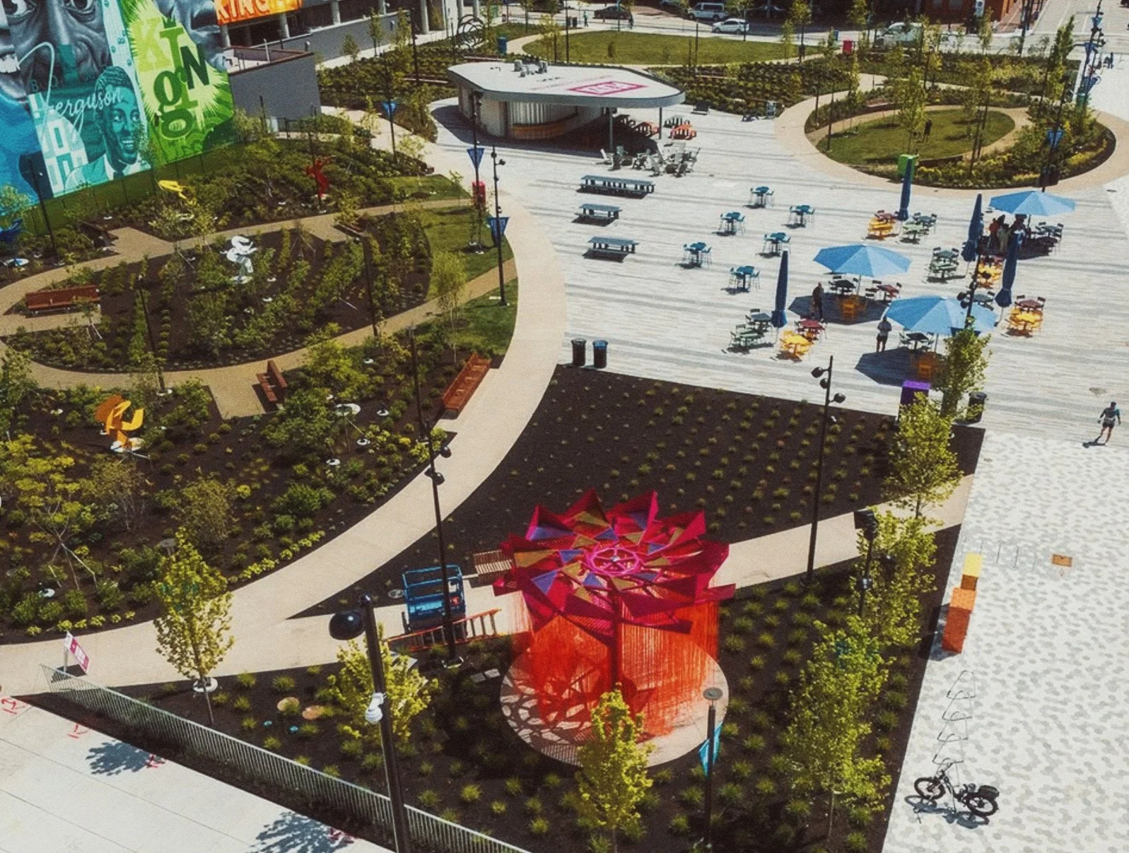 Aerial view of a public park with landscaped garden areas, pathways, benches, trees, and a seating area with blue umbrellas. There is a stage or pavilion in the center with people around, and colorful sculptures and structures throughout the park.