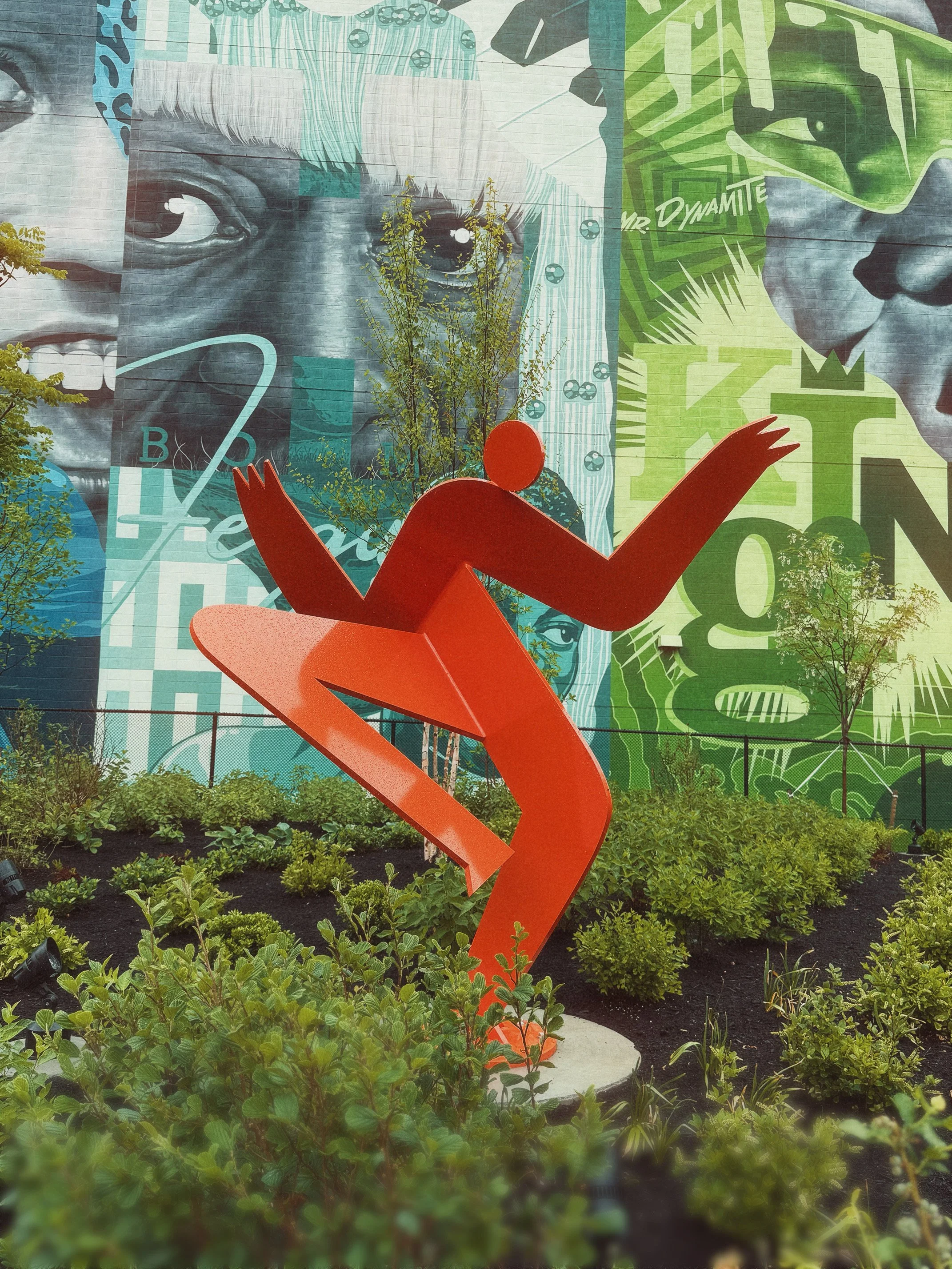 Colorful mural featuring two faces, one with a headband with blue spots, and the other with sunglasses, surrounded by vibrant green and blue patterns. In the foreground, an orange abstract sculpture of a person sitting with one arm raised, set in a garden with small green plants and two small trees.