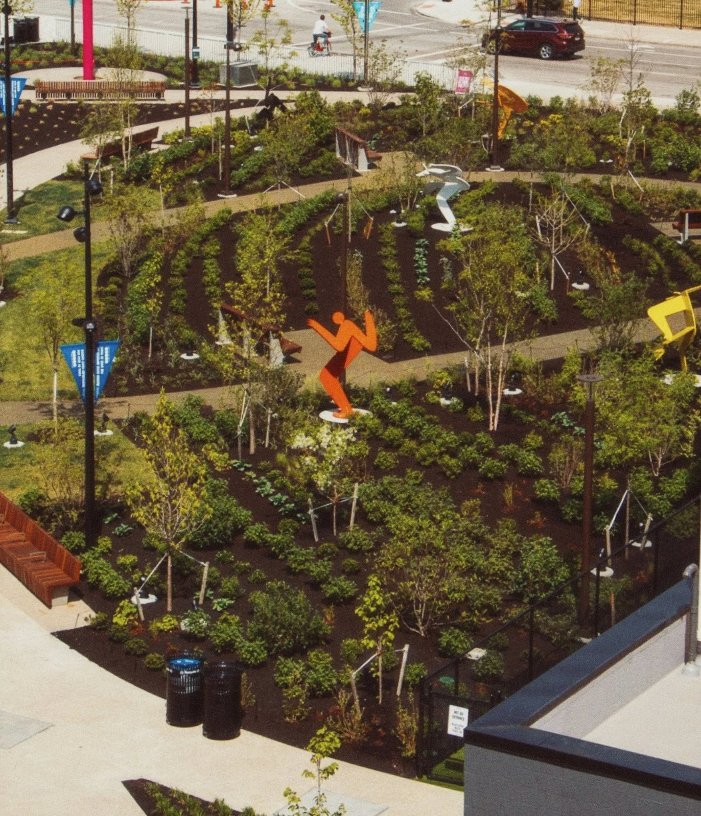 A rooftop garden with trees, bushes, and small plants, featuring colorful sculptures of a person and a bird. Nearby are benches, pathways, and a street with cars and a cyclist in the background.