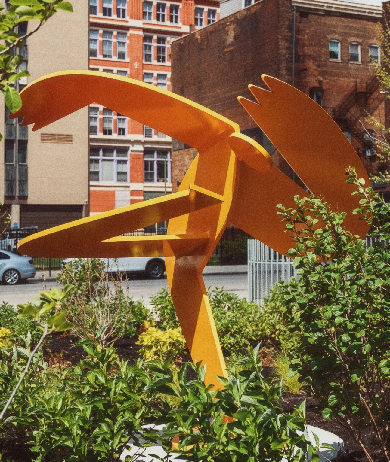 A large, abstract, orange sculpture resembling a bird, placed among green bushes in an urban setting with buildings and parked cars in the background.