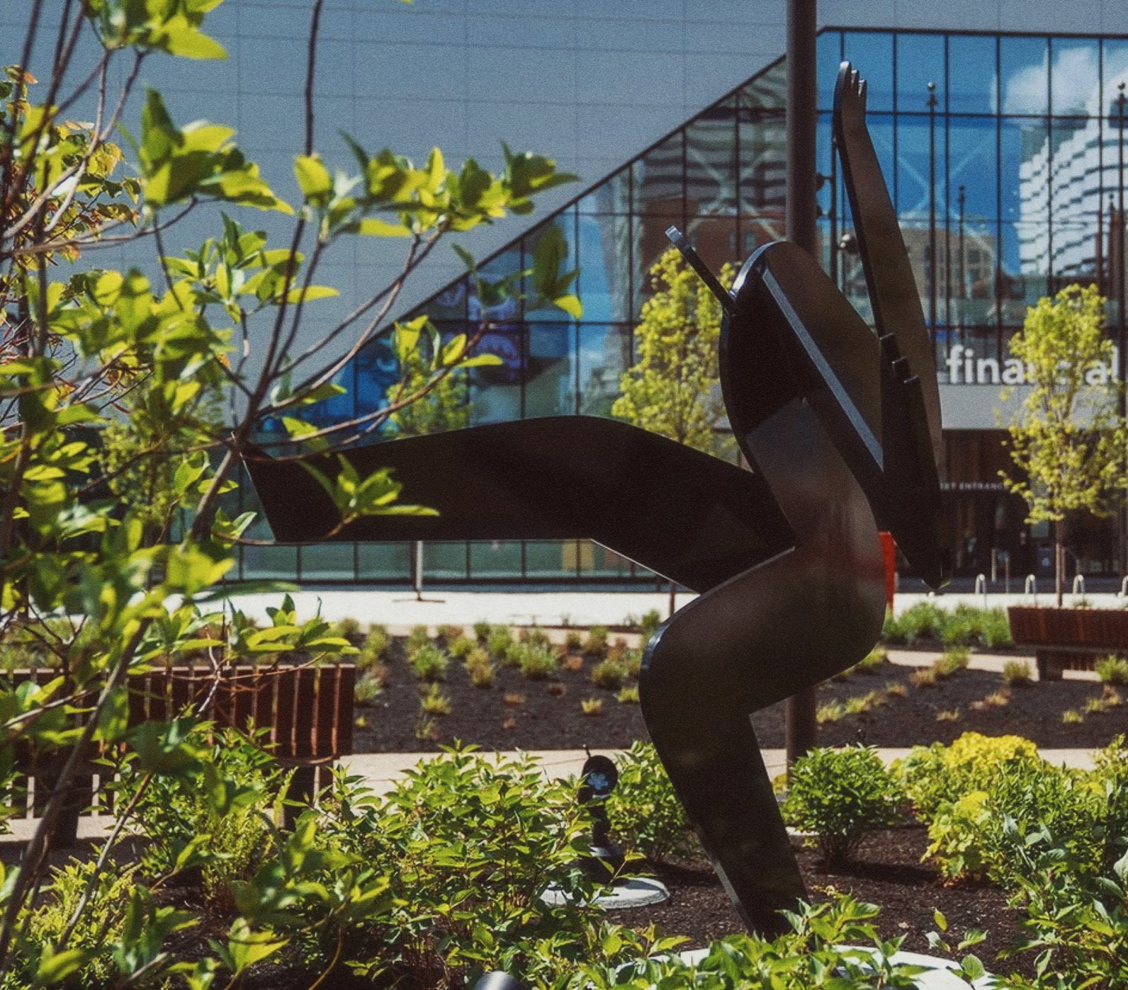 A modern black sculpture of an angel with large wings situated in a landscaped urban park with green shrubbery, trees, and a glass building in the background.