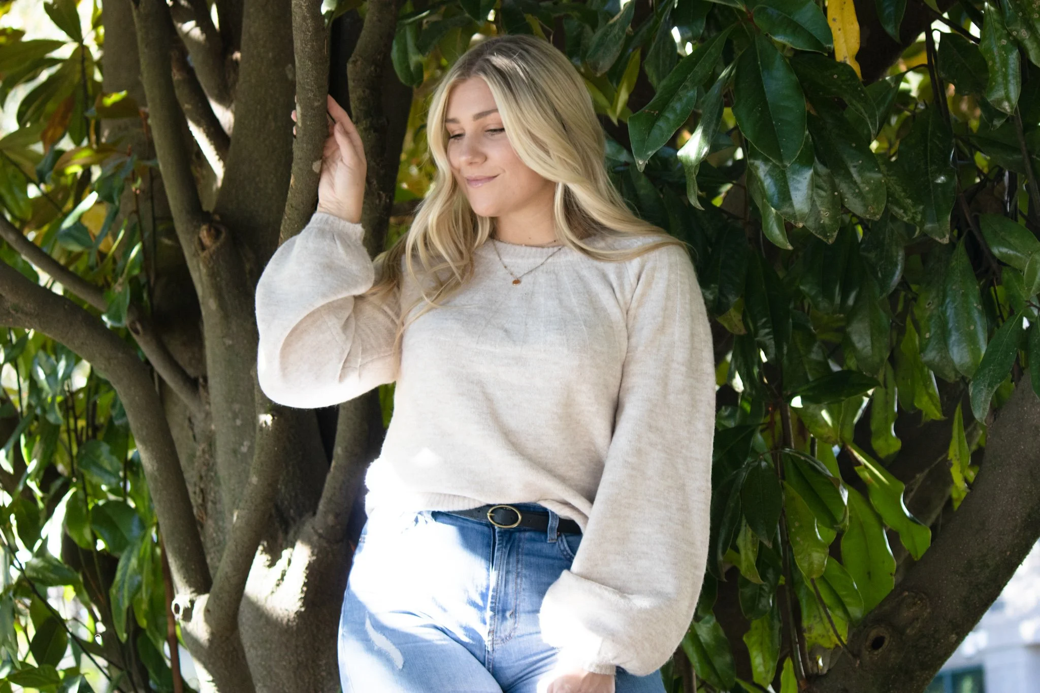 Young woman with long blonde hair, wearing a beige sweater and light blue jeans, leaning on a tree branch outdoors with green leaves.
